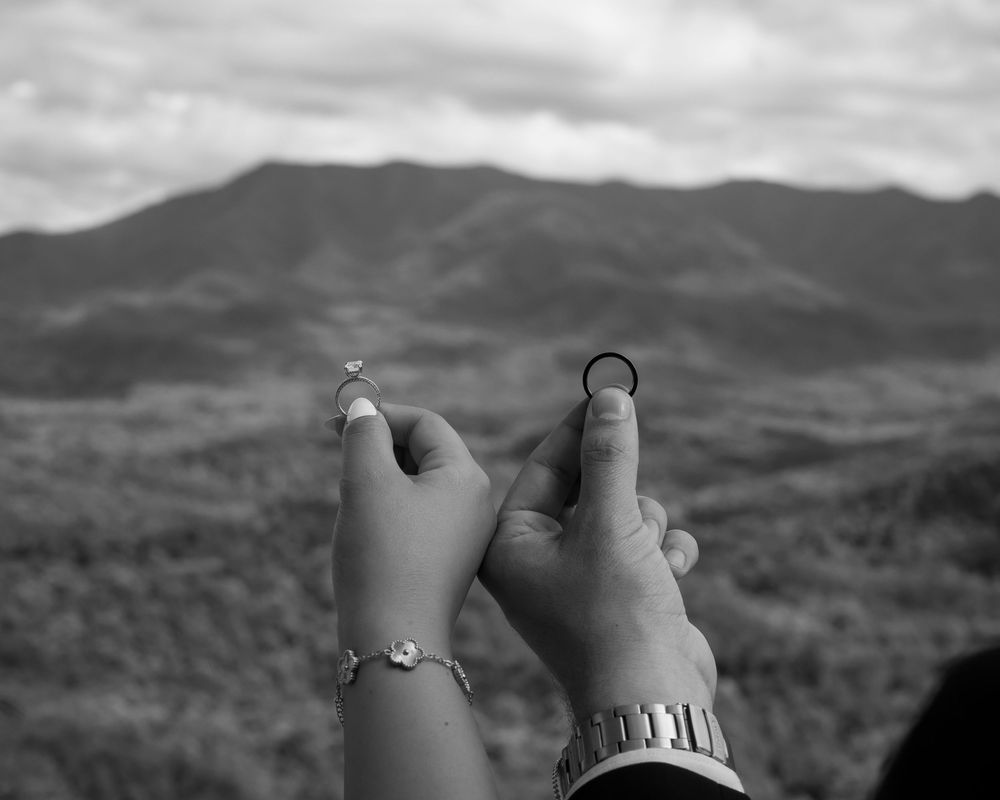 Black and white photo of couple holding wedding rings with mountain backdrop and dramatic cloudy sky.