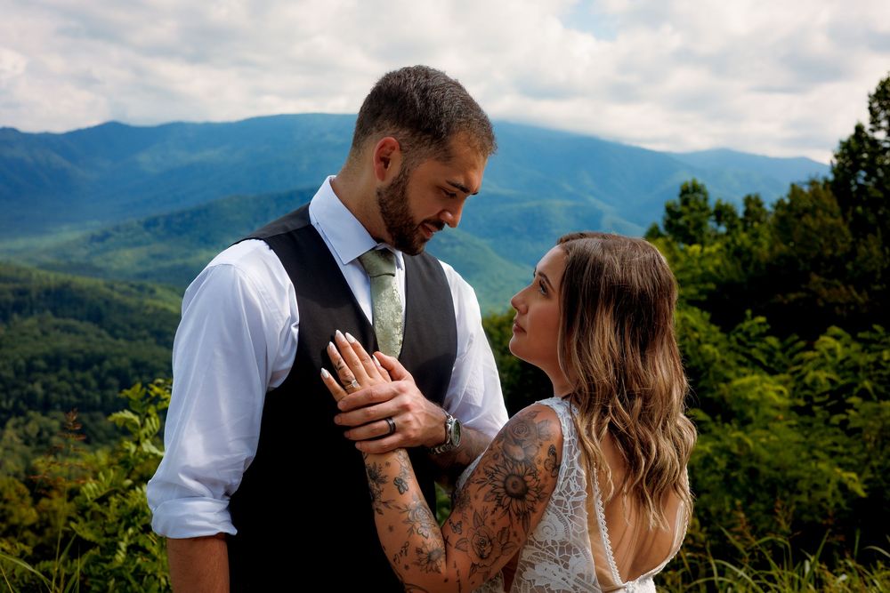Couple in wedding attire sharing romantic moment with scenic mountain backdrop and lush green trees.