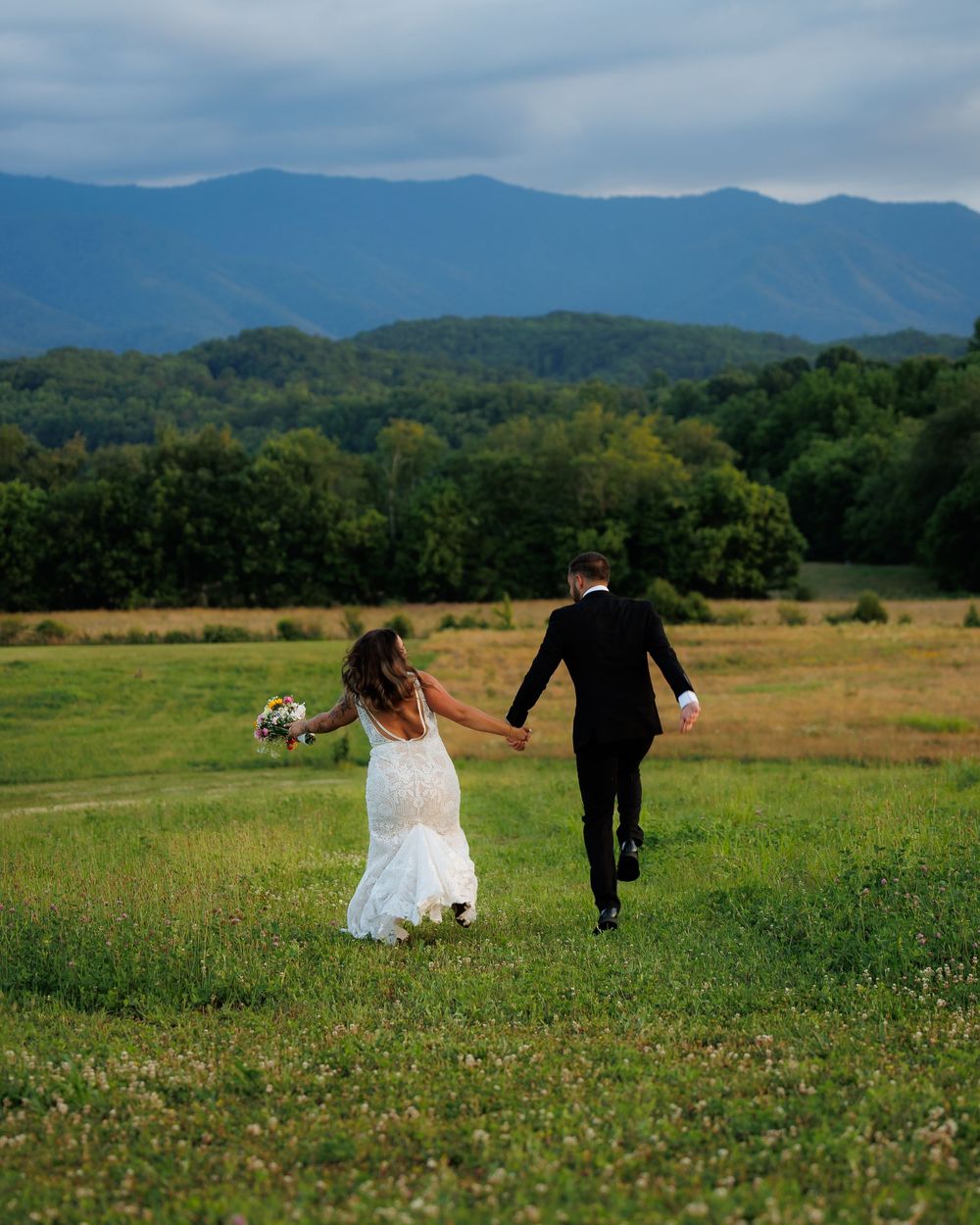 Newlyweds walk hand-in-hand through green meadow with stunning Blue Ridge Mountains behind.