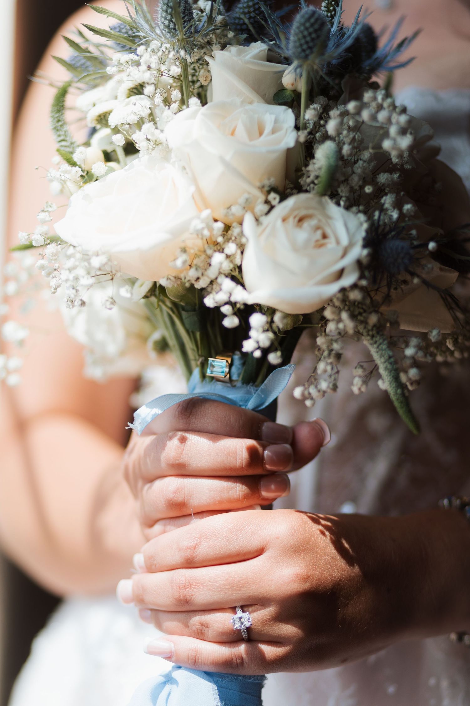 Bride holding a wedding bouquet of white roses, blue thistles, and baby's breath, wearing a diamond engagement ring.