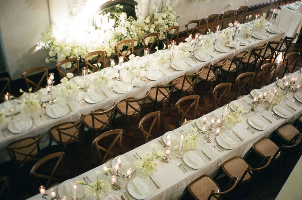Elegant wedding reception with long banquet tables, white floral centerpieces, candles, and cross-back chairs in a bright venue.