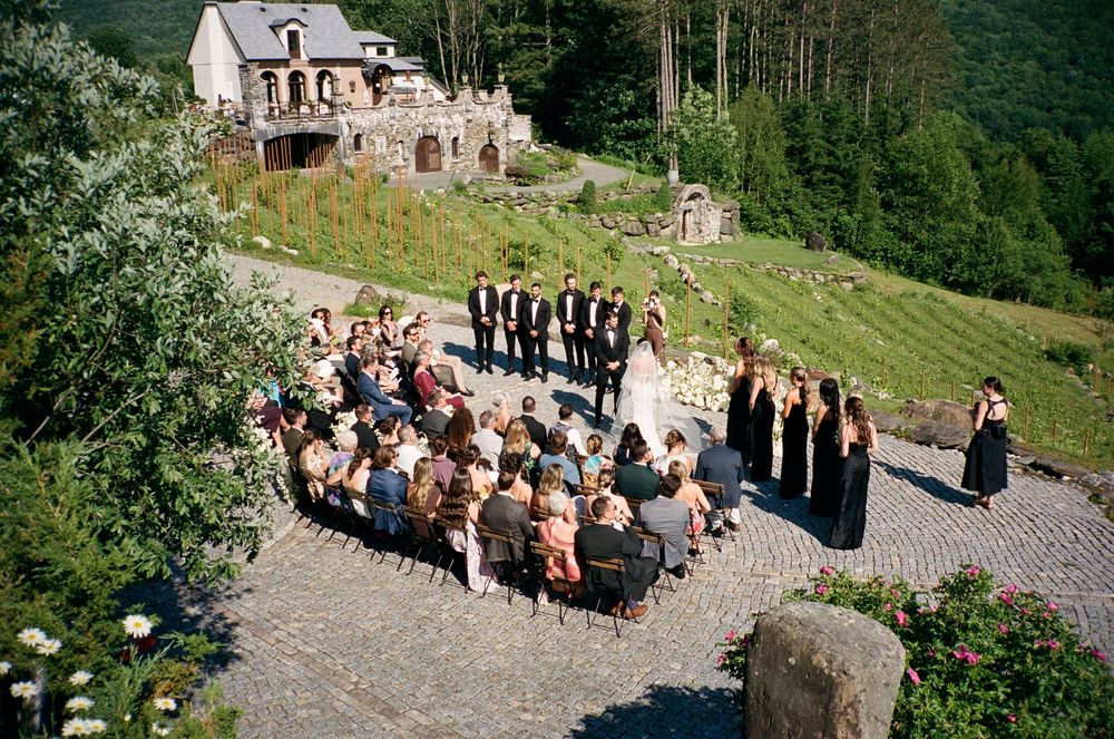 Outdoor wedding ceremony on cobblestone terrace with guests seated, bridal party in black, and rustic stone ruins backdrop.