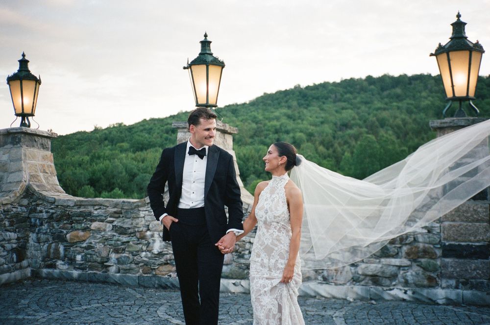 Bride and groom share a romantic moment on stone terrace, veil flowing in wind, lush green hills at dusk.