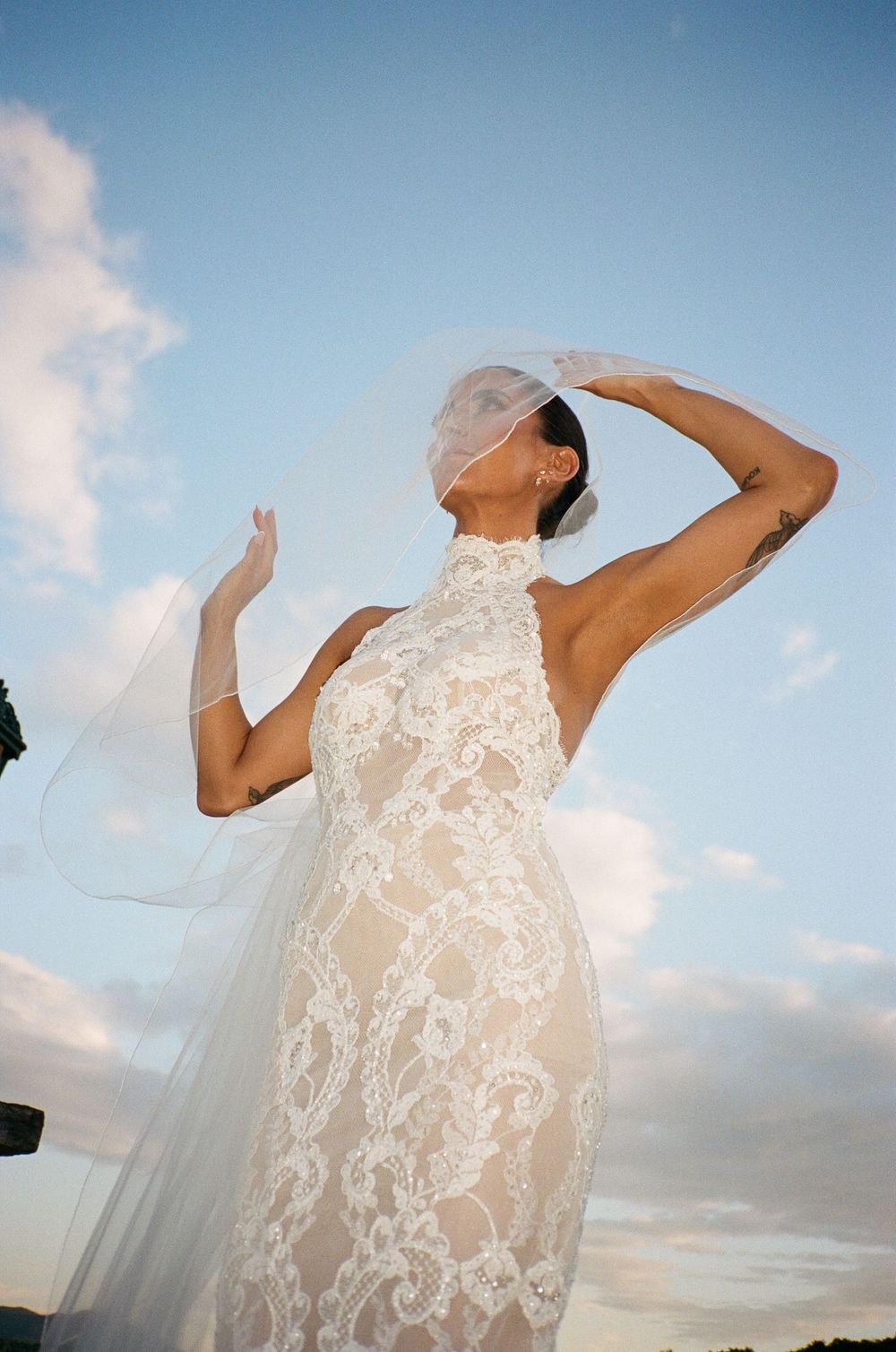 Bride in elegant halter lace wedding gown with flowing veil poses against blue sky at golden hour.
