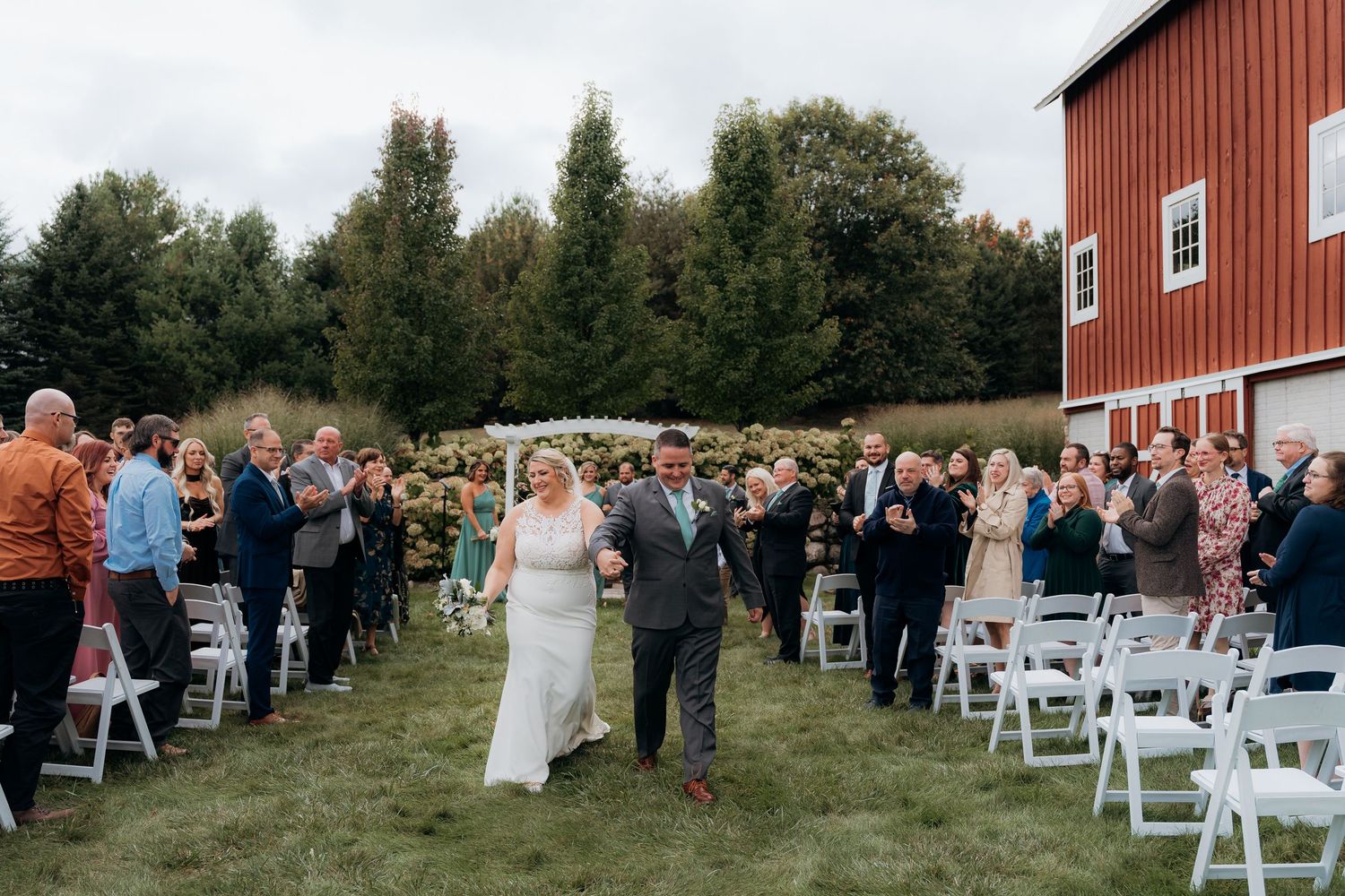 Bride and groom walk down outdoor aisle at barn wedding venue, surrounded by cheering guests on a lush green lawn.