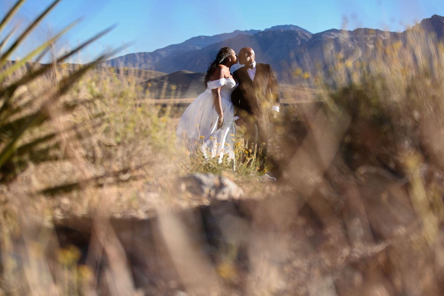 A bride and groom embrace in a desert landscape with mountains and blue sky behind them.