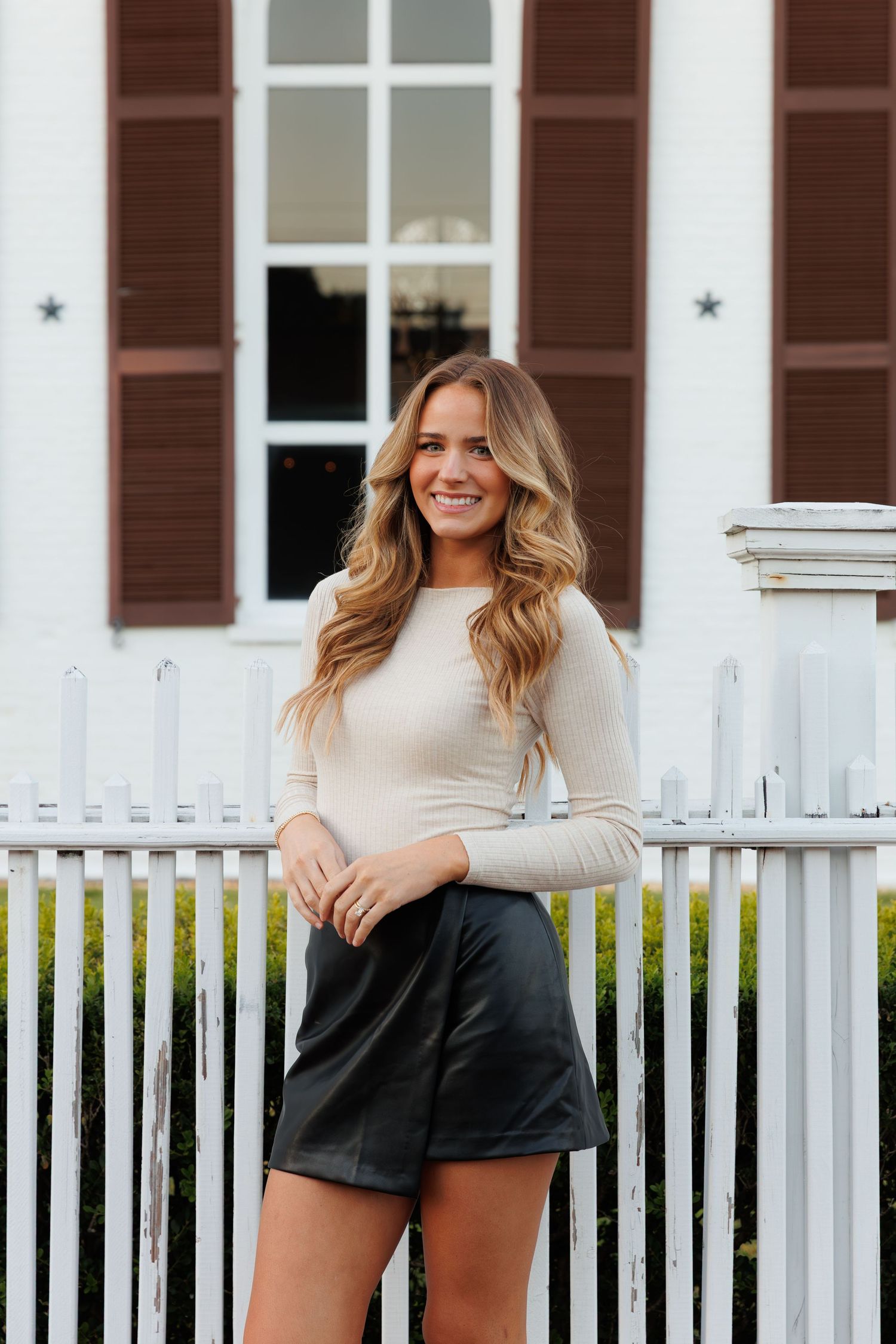 Woman in cream top and black mini skirt smiling by white picket fence near arched windows.