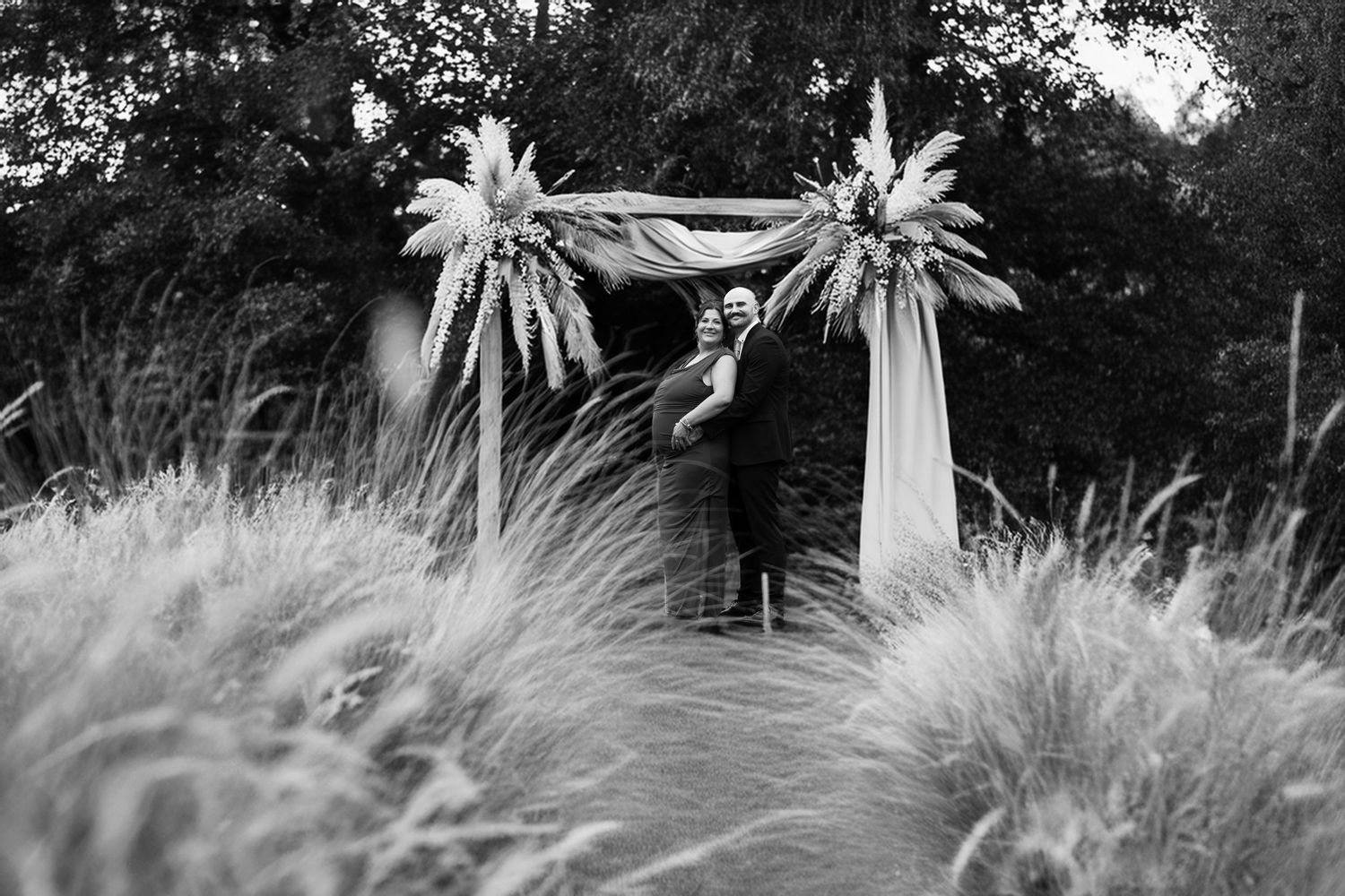 Black and white photo of a person standing under a floral arch with palm leaves and draped fabric outdoors.