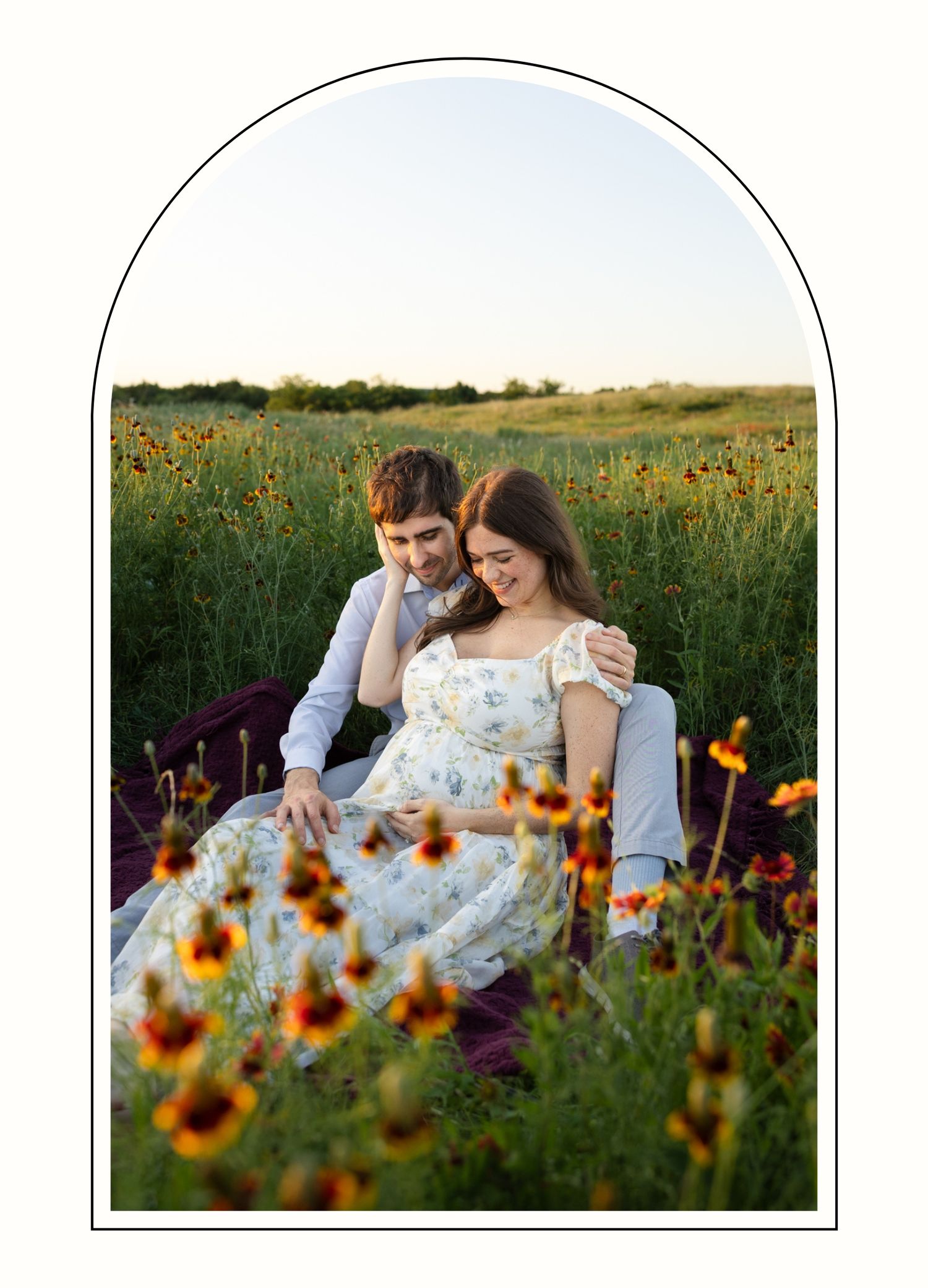 Pregnant couple sitting and cuddling in a field of red and orange flowers smiling at her baby bump