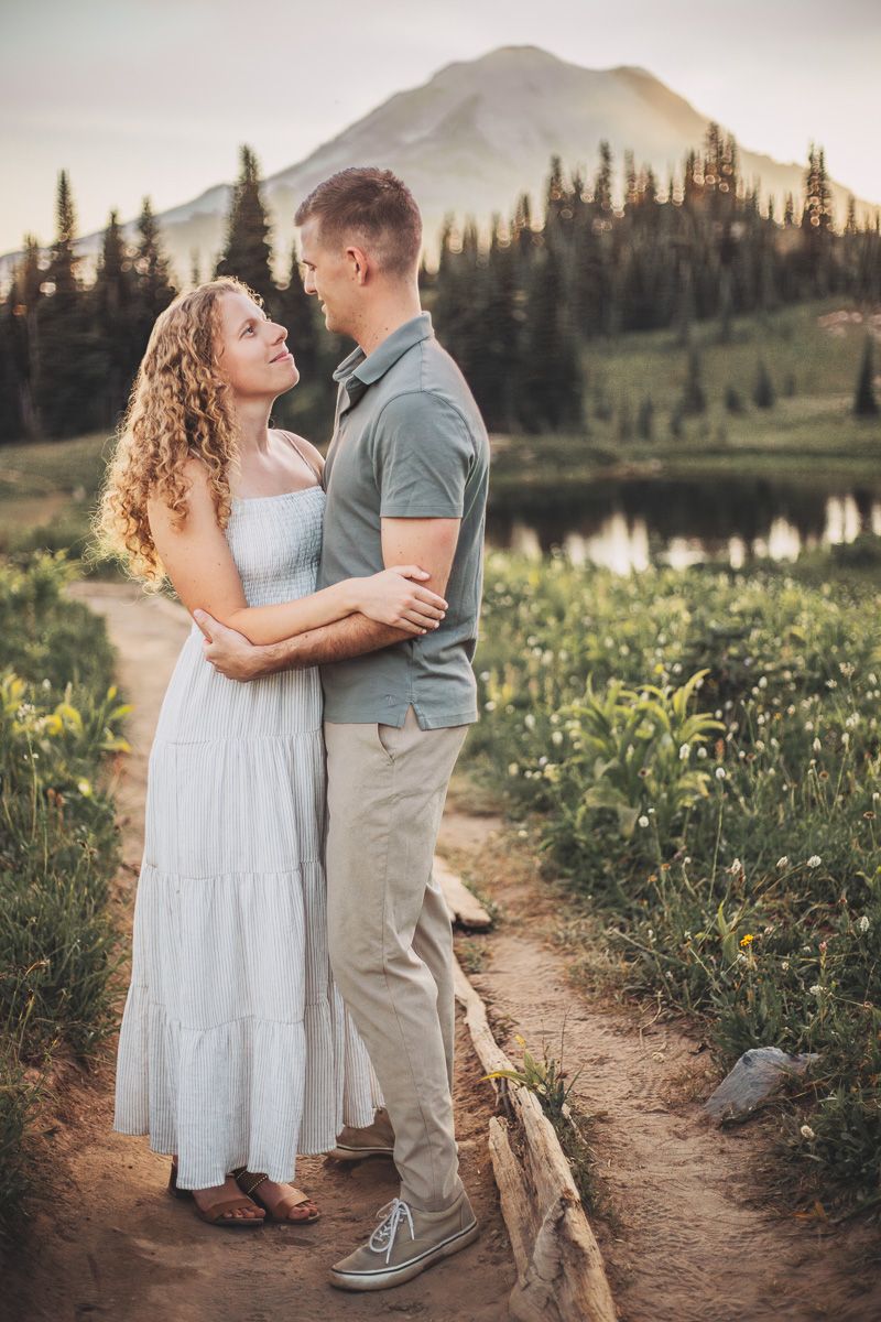 Couple holding each other on a mountain path beside a lake with snow-capped Mount Rainier backdrop.