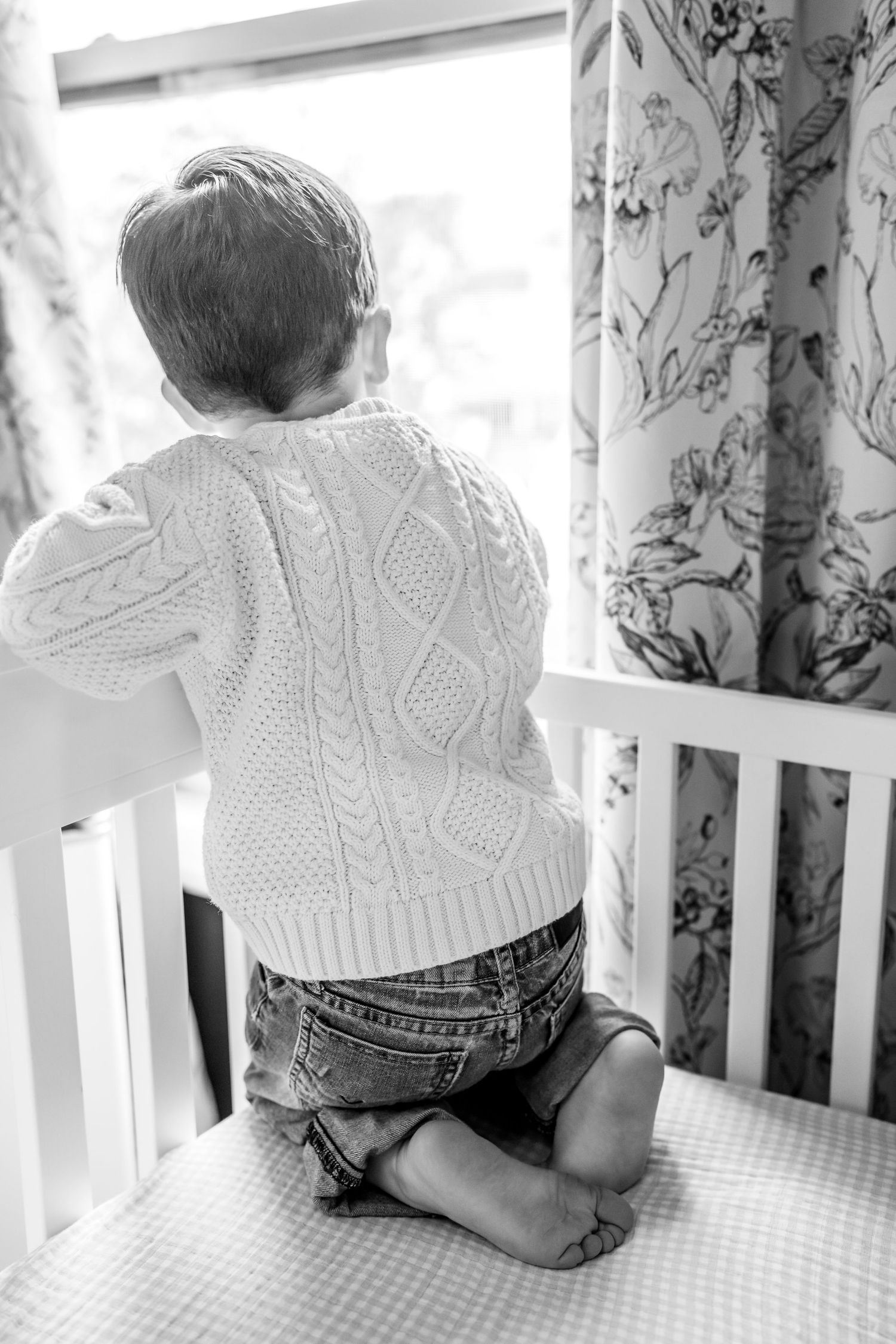 Black and white photo of a toddler in a cable-knit sweater and jeans kneeling in a white crib, looking out a window at a lifestyle session.