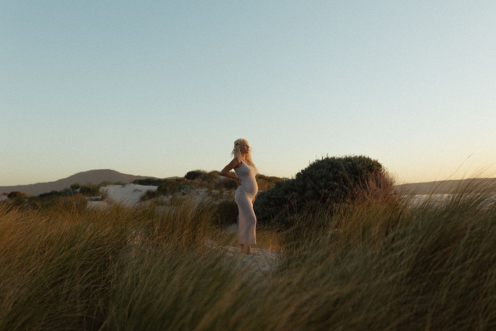 Pregnant woman standing in coastal sand dunes at golden hour, surrounded by tall grass with ocean views.