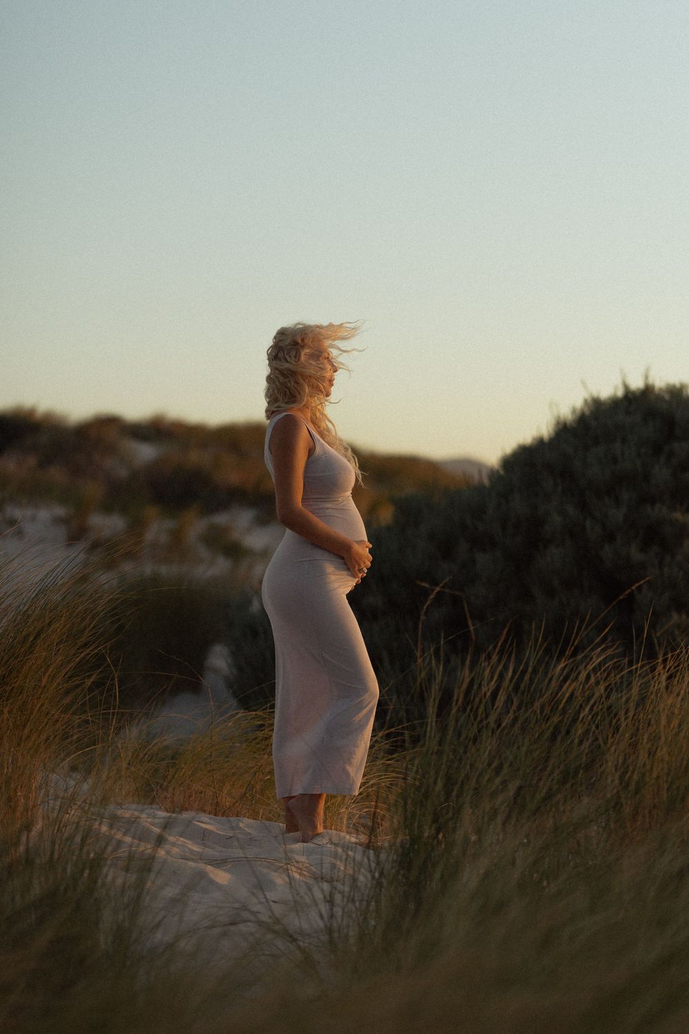 Pregnant woman in white dress standing on sandy dunes at golden hour.