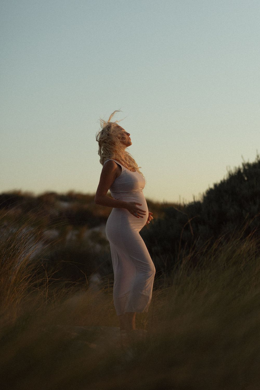 Pregnant woman in white dress standing in golden hour light among grass dunes, hair blowing in the wind.