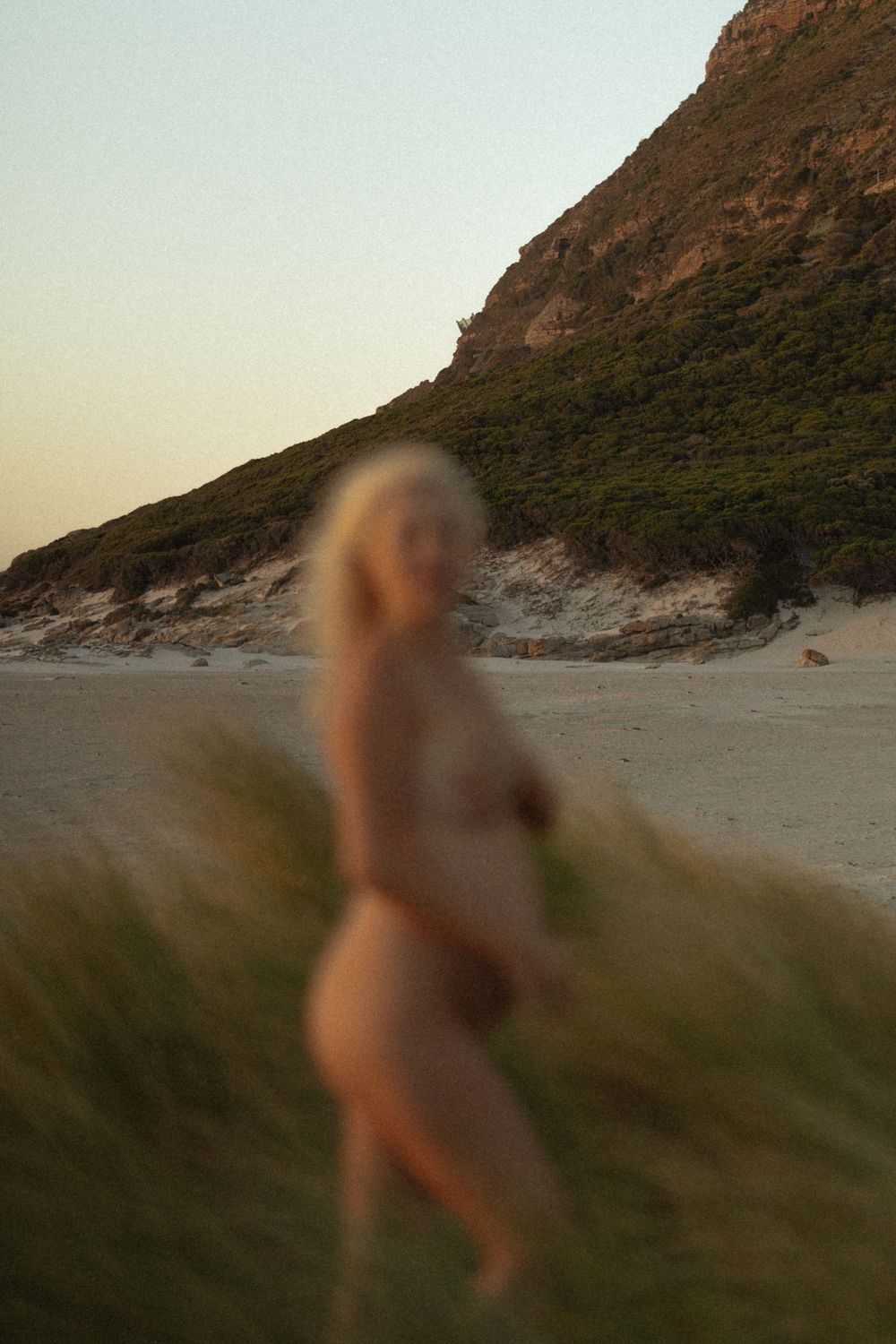Blurred artistic shot of nude pregnant woman standing in beach grass dunes near rocky hill.