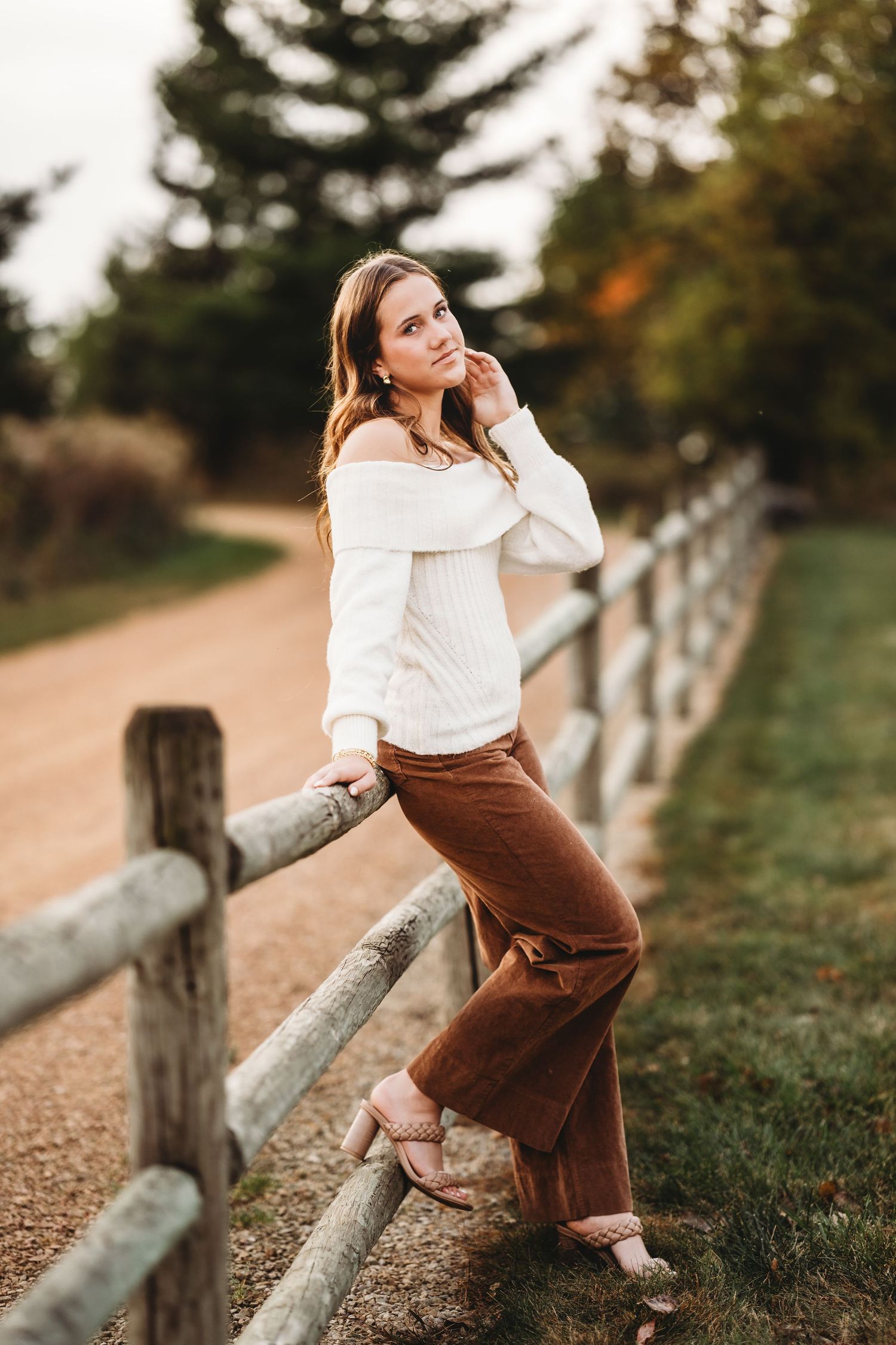Young woman in white sweater and brown velvet pants poses by rustic wooden fence on a scenic autumn path.
