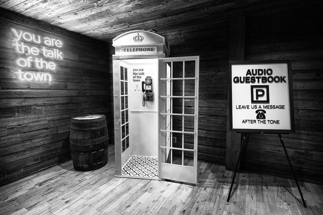A black and white photo shows a vintage British telephone booth with neon text and wooden walls in a dark interior space.