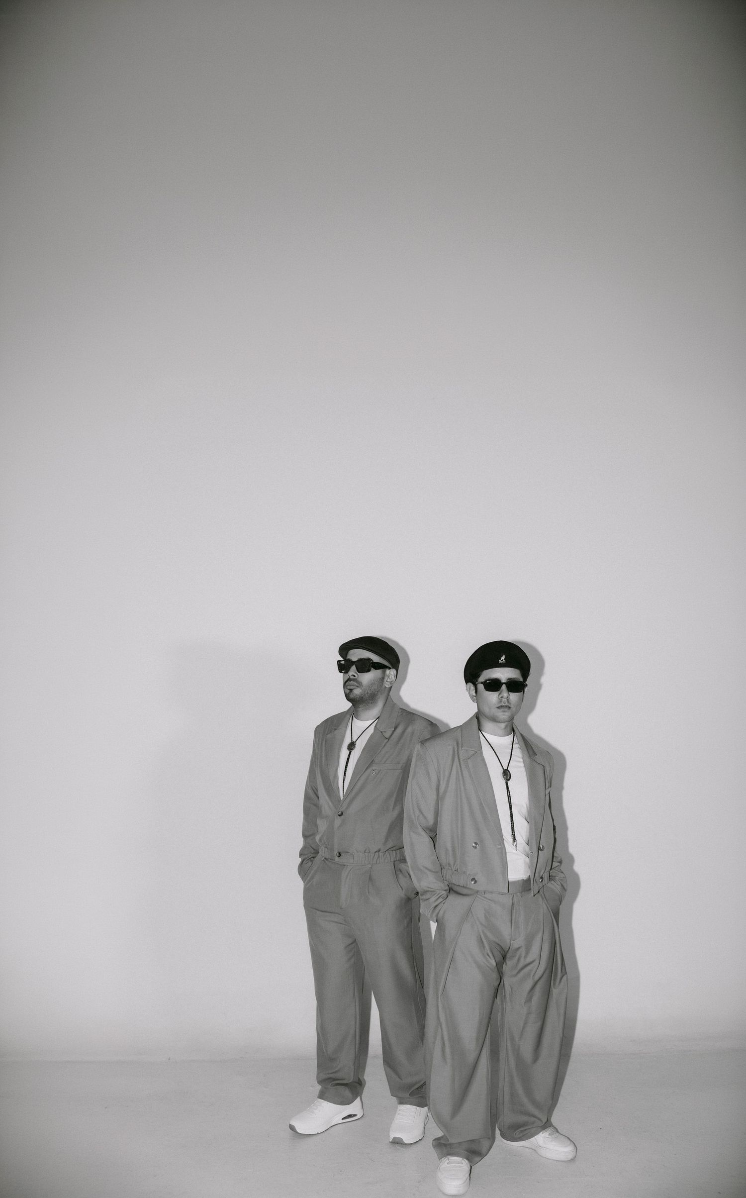 Two men in matching gray suits, flat caps, and sunglasses pose together in a black and white studio photo.