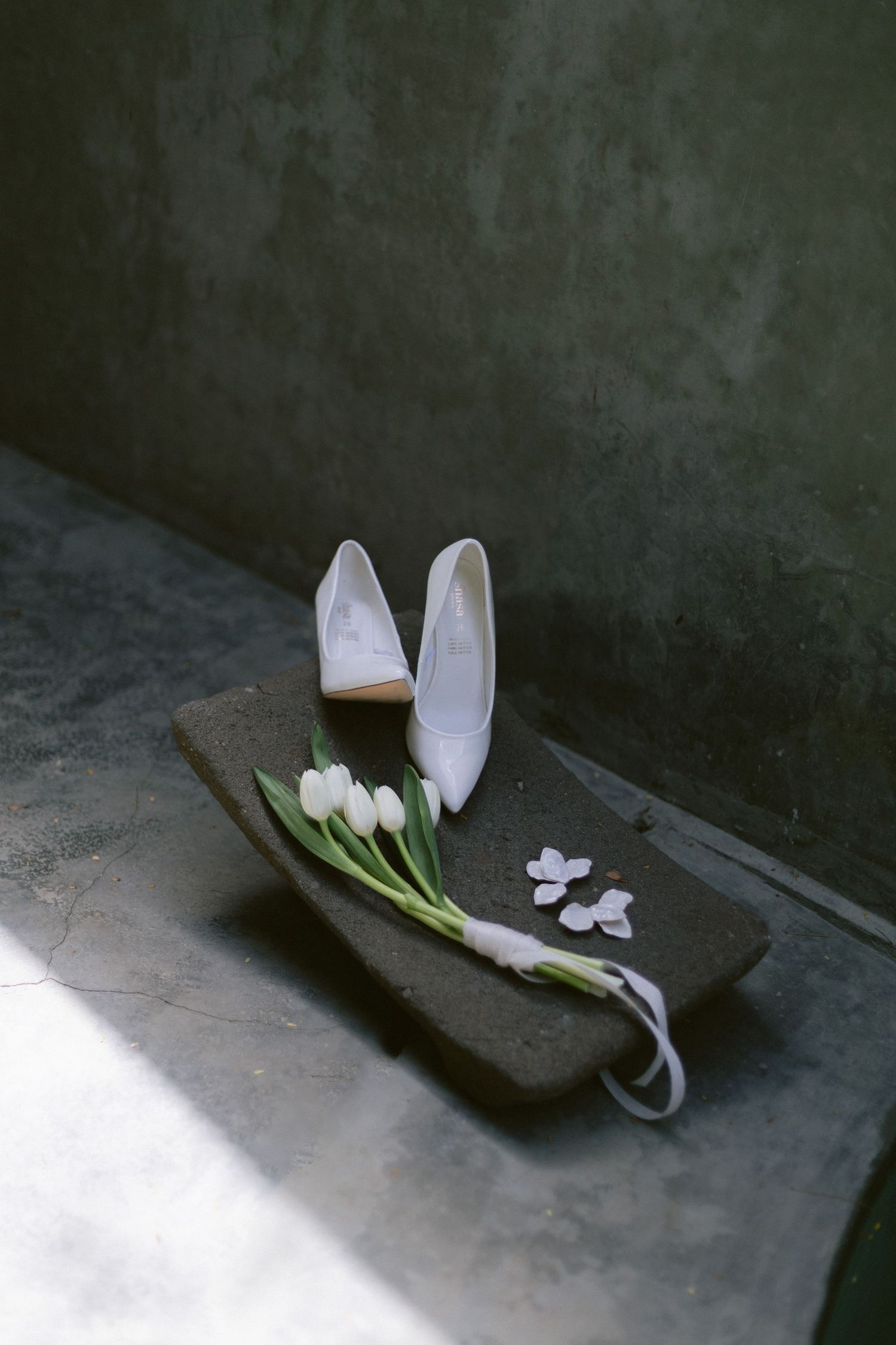White bridal heels and white tulip bouquet arranged on a dark stone surface in moody natural light.