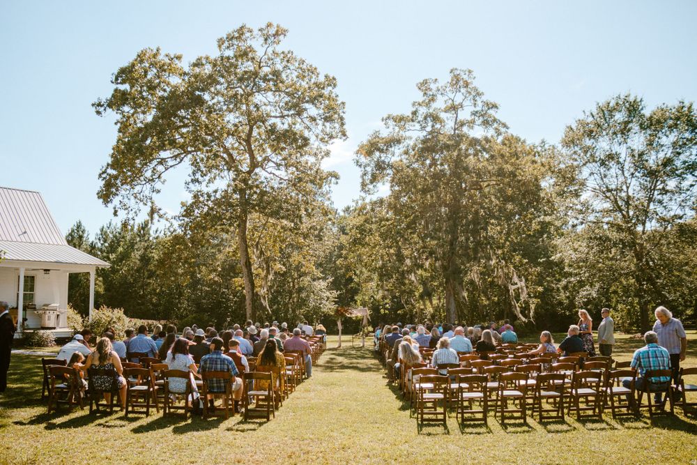 Outdoor wedding ceremony setup with wooden chairs and guests under large oak trees.