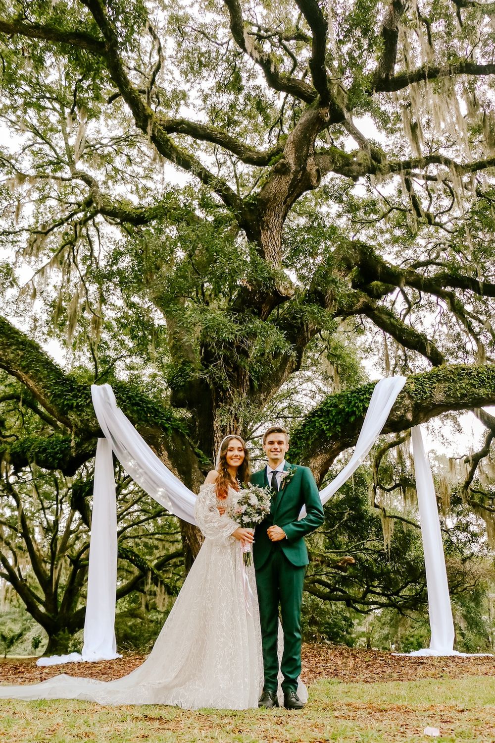 Newlyweds pose under white draped arch and ancient oak tree in elegant garden setting.