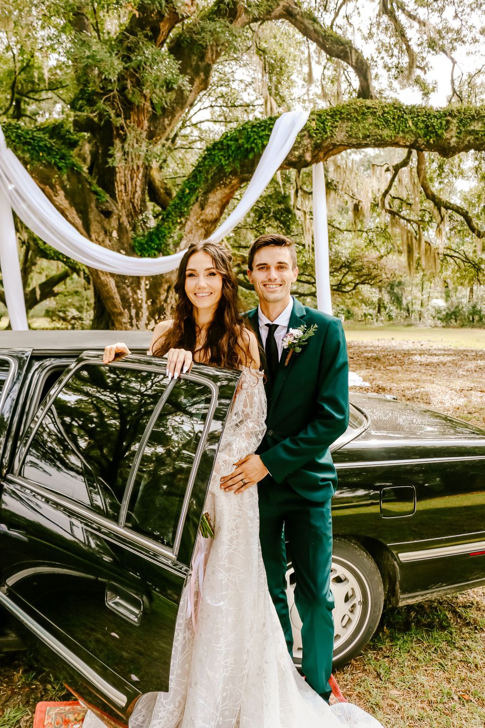 Newlyweds pose by vintage black car under trees draped with white fabric.
