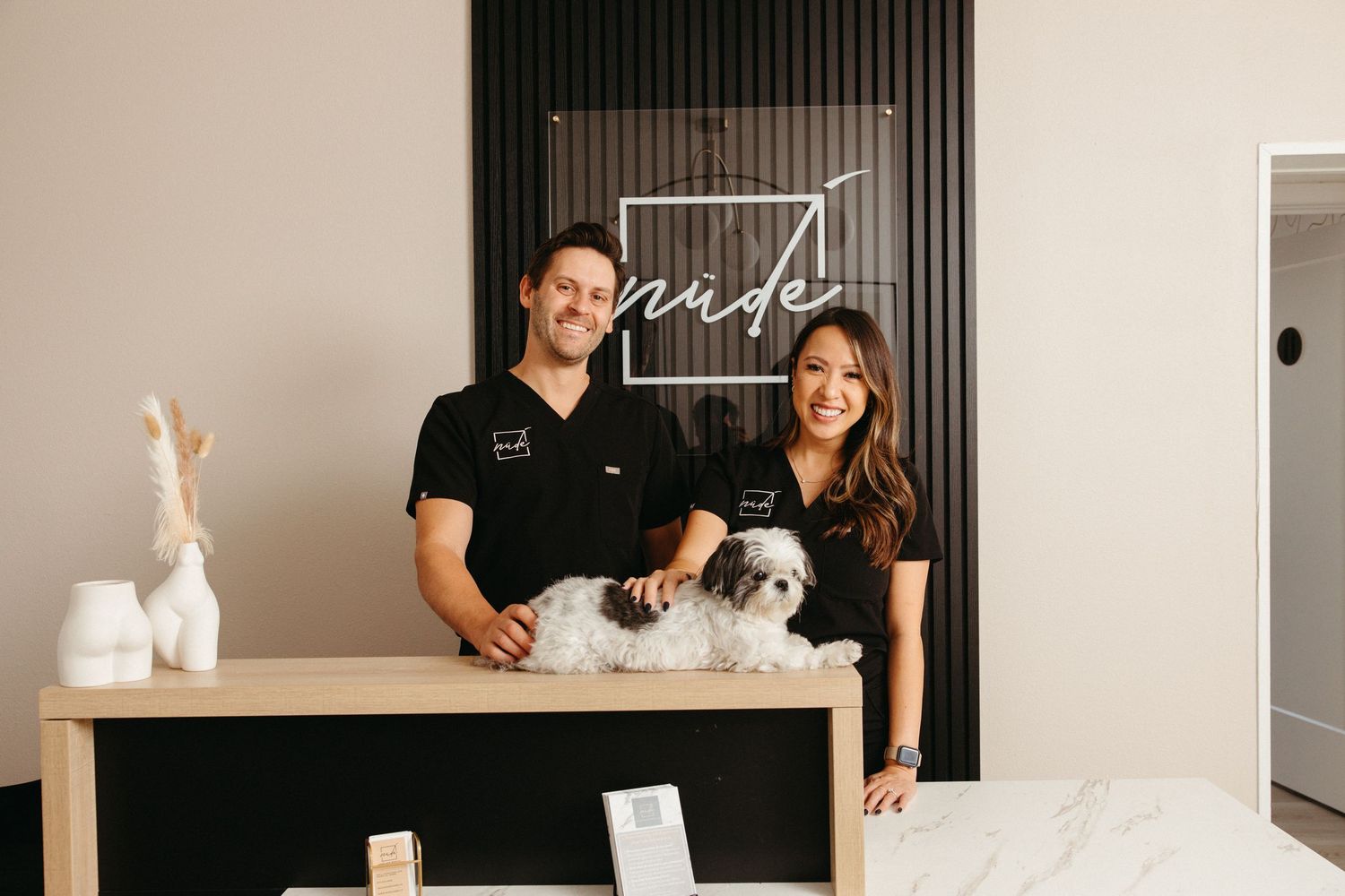 A man and woman smile behind a reception desk with a small dog at a modern salon called Nude.