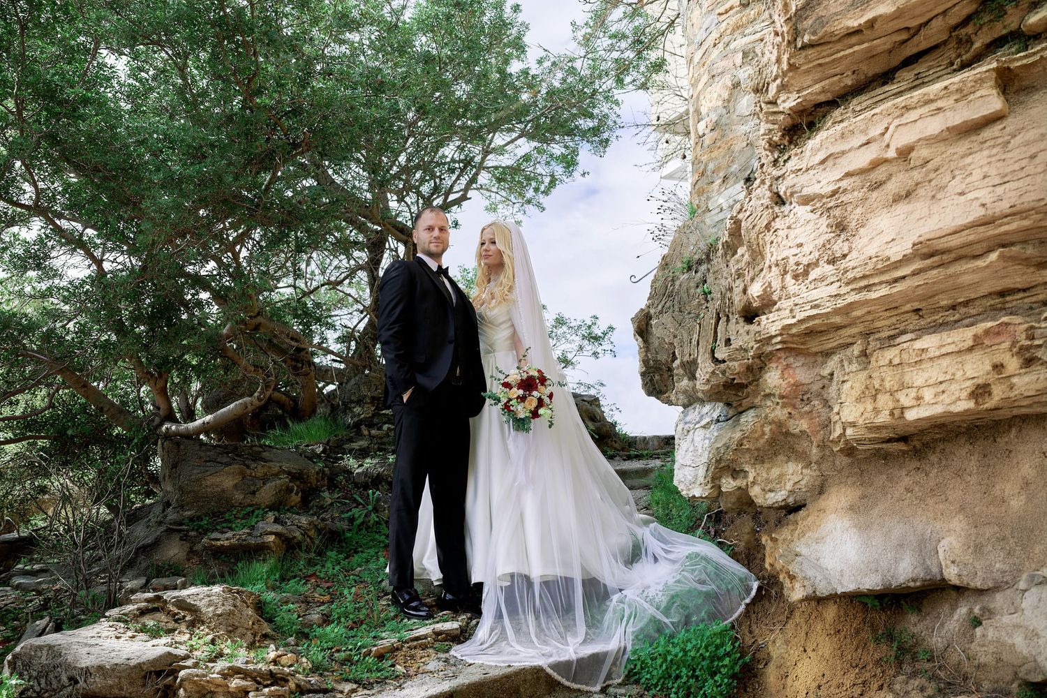 Full-length portrait of a wedding couple with a large veil near rocks and trees.