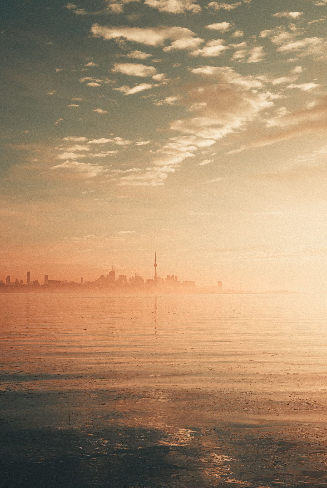 Hazy golden sunrise over Toronto city skyline reflected on calm Lake Ontario waters.