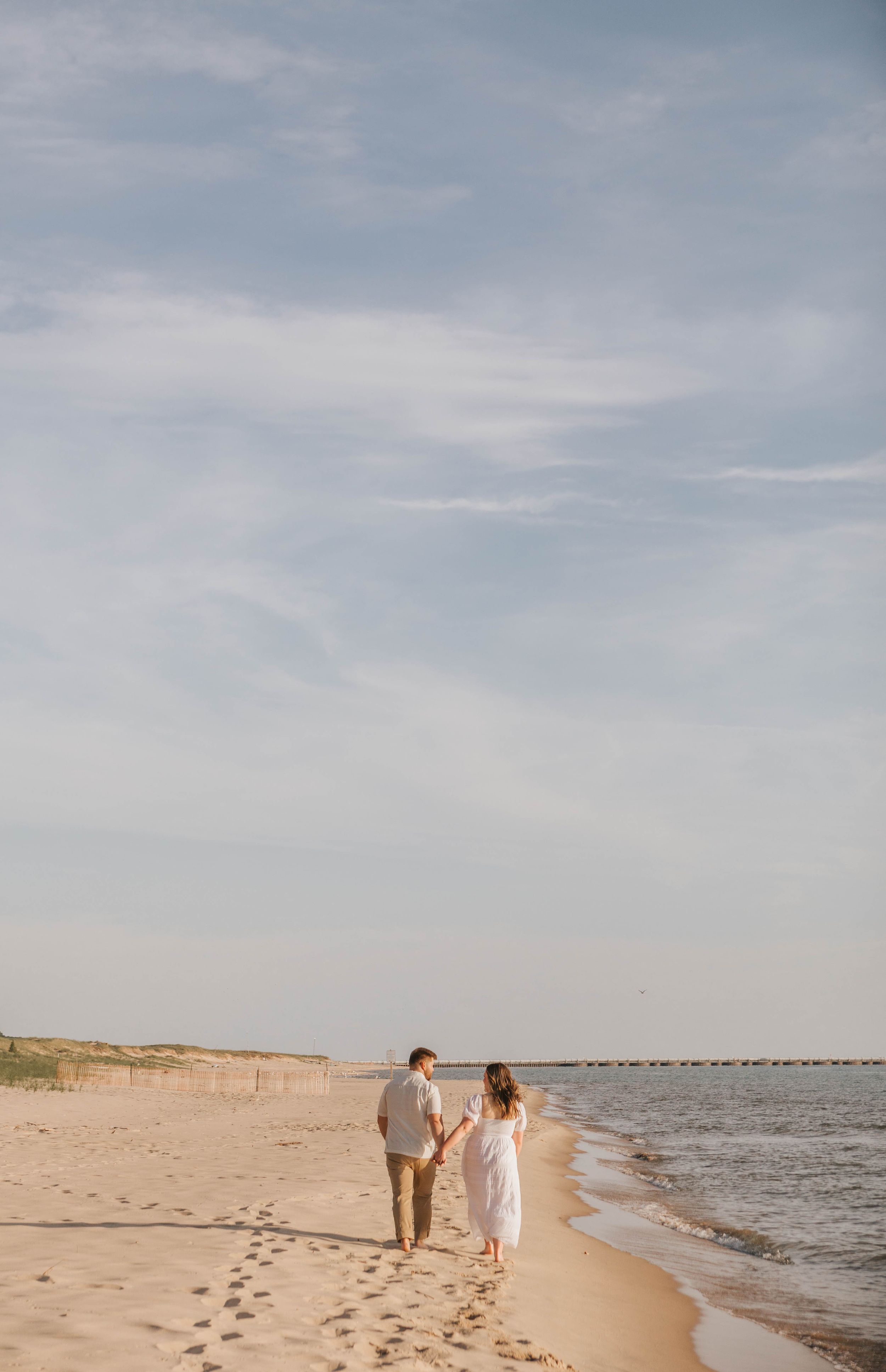 A couple walks hand-in-hand along a sandy beach shoreline under a soft cloudy sky.