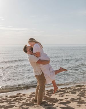 A couple shares a romantic kiss on a sandy beach, as the man lifts the woman in a white dress at sunset.