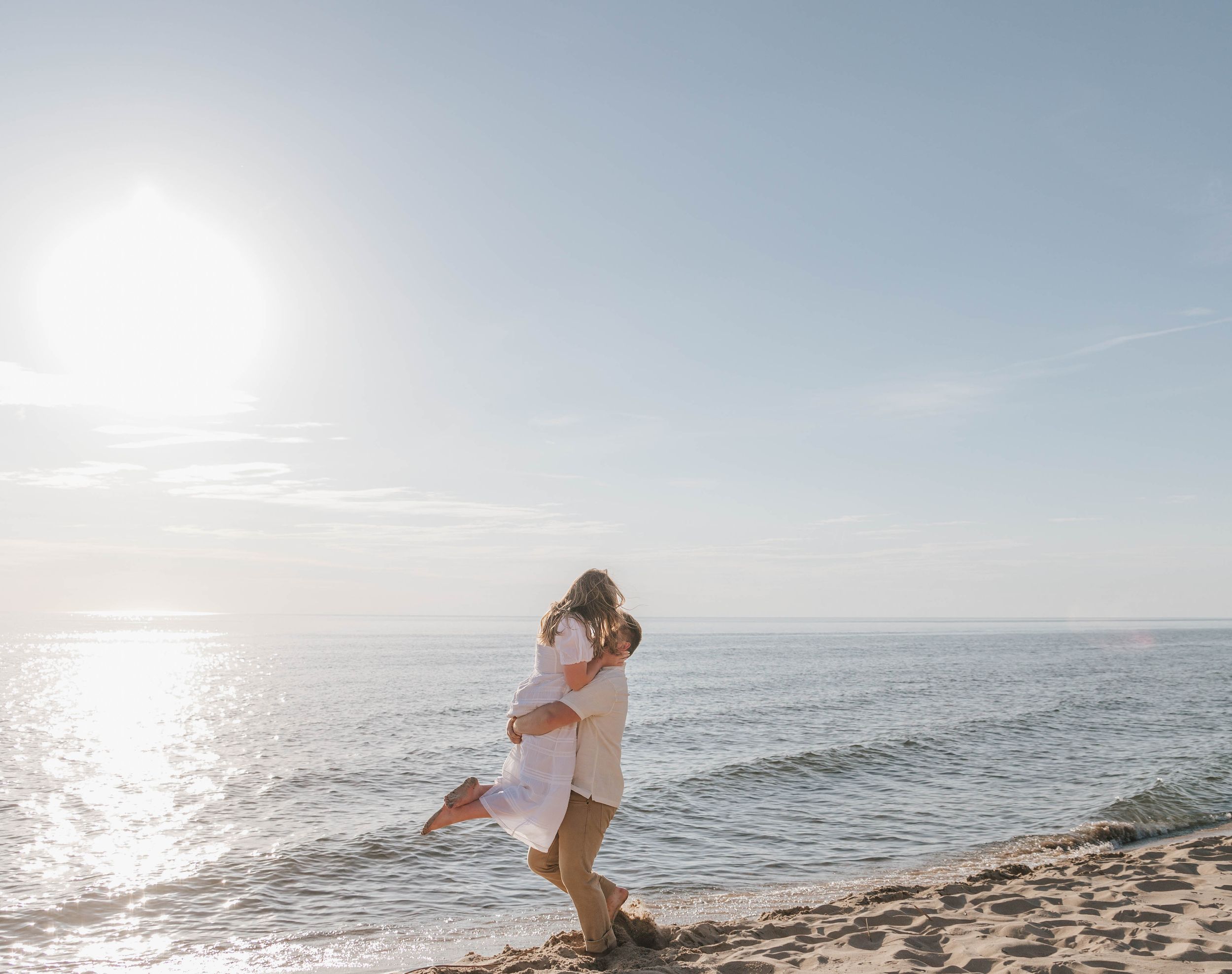 Couple embracing on a sunny beach, man lifting woman in romantic pose with ocean and bright sky backdrop.