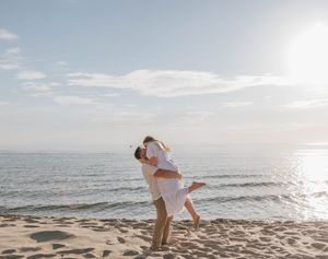 Couple embracing and kissing on a sunny beach, man lifting woman in white dress near ocean shoreline.