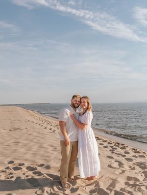 A couple embraces on a sandy beach, the woman in a white dress and man in a white shirt, with calm ocean waters behind them.