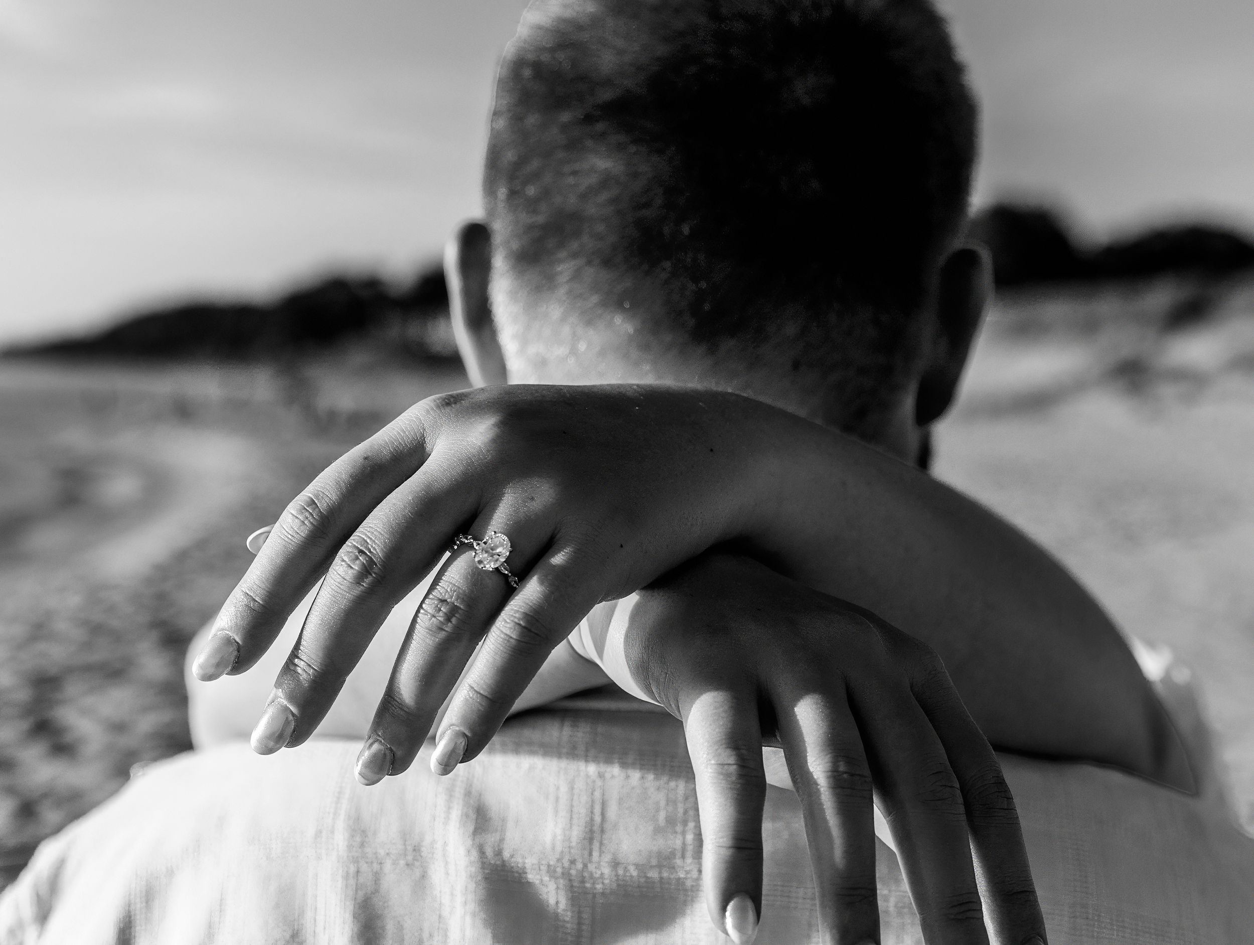 Black and white photo of couple embracing on beach, woman's hand showing diamond engagement ring on man's shoulder.
