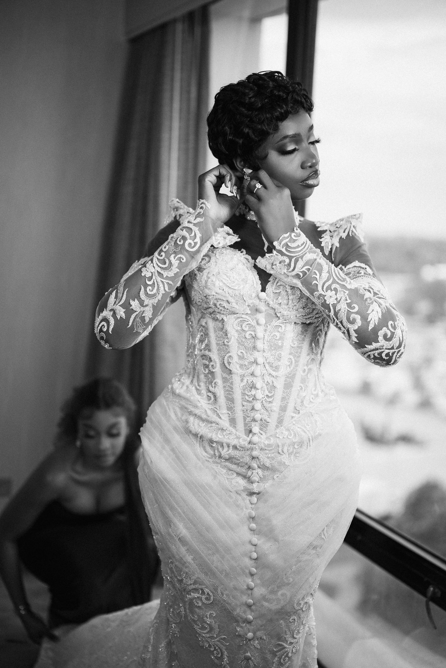 Black and white photo of a bride in a lace mermaid wedding gown putting on earrings near a window.
