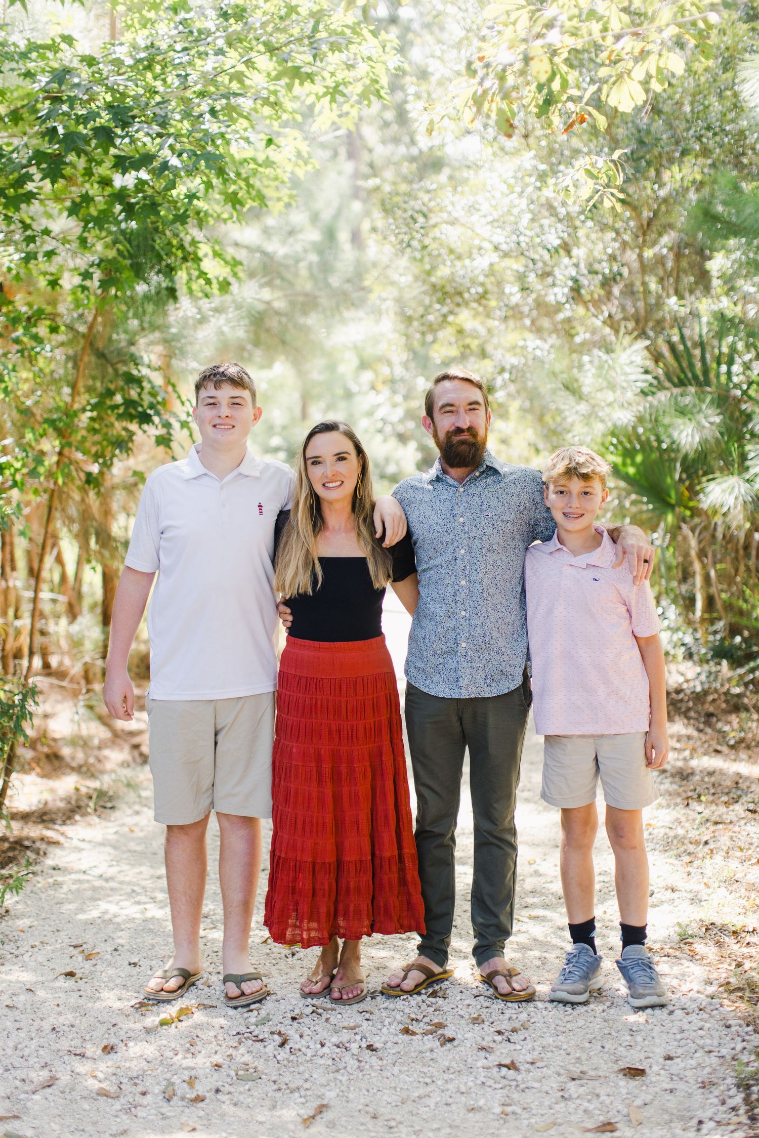 Family of four posing on a sunlit garden path surrounded by lush green trees, wearing casual summer outfits in a Hilton Head, SC park.