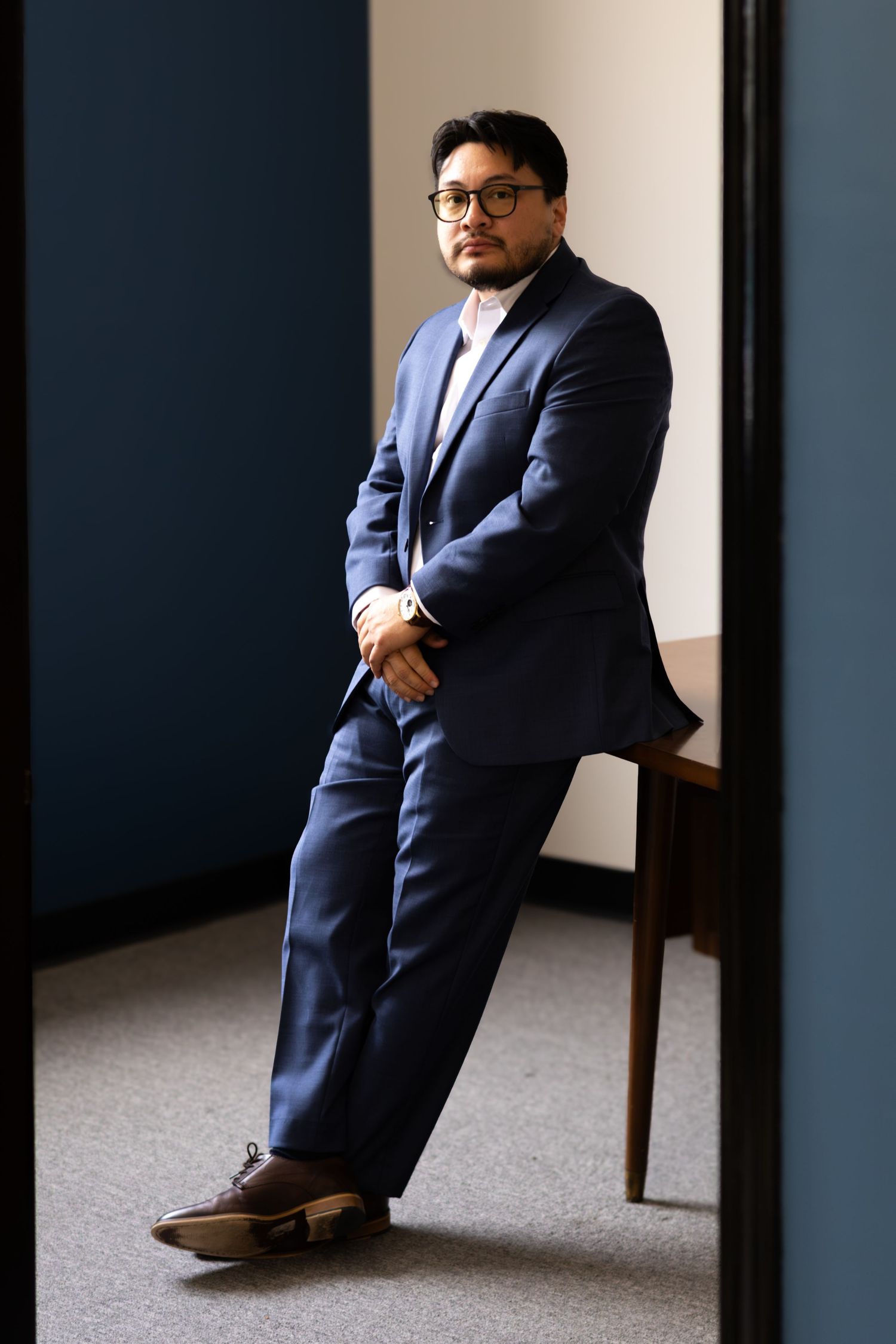 Professional man in blue suit and glasses leaning against wall in modern office setting.