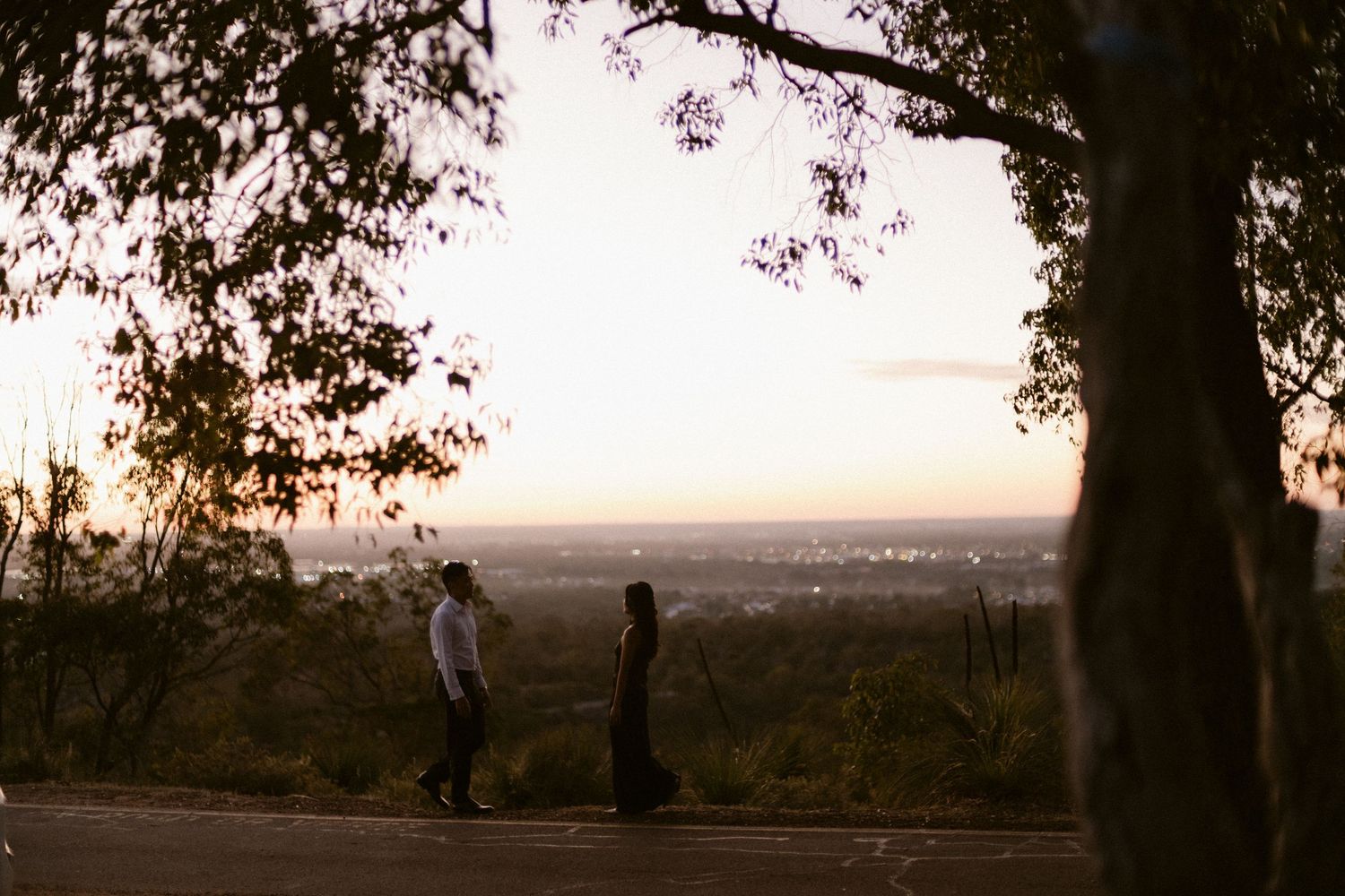 Silhouette of couple standing apart on hilltop road at sunset with city skyline in background.