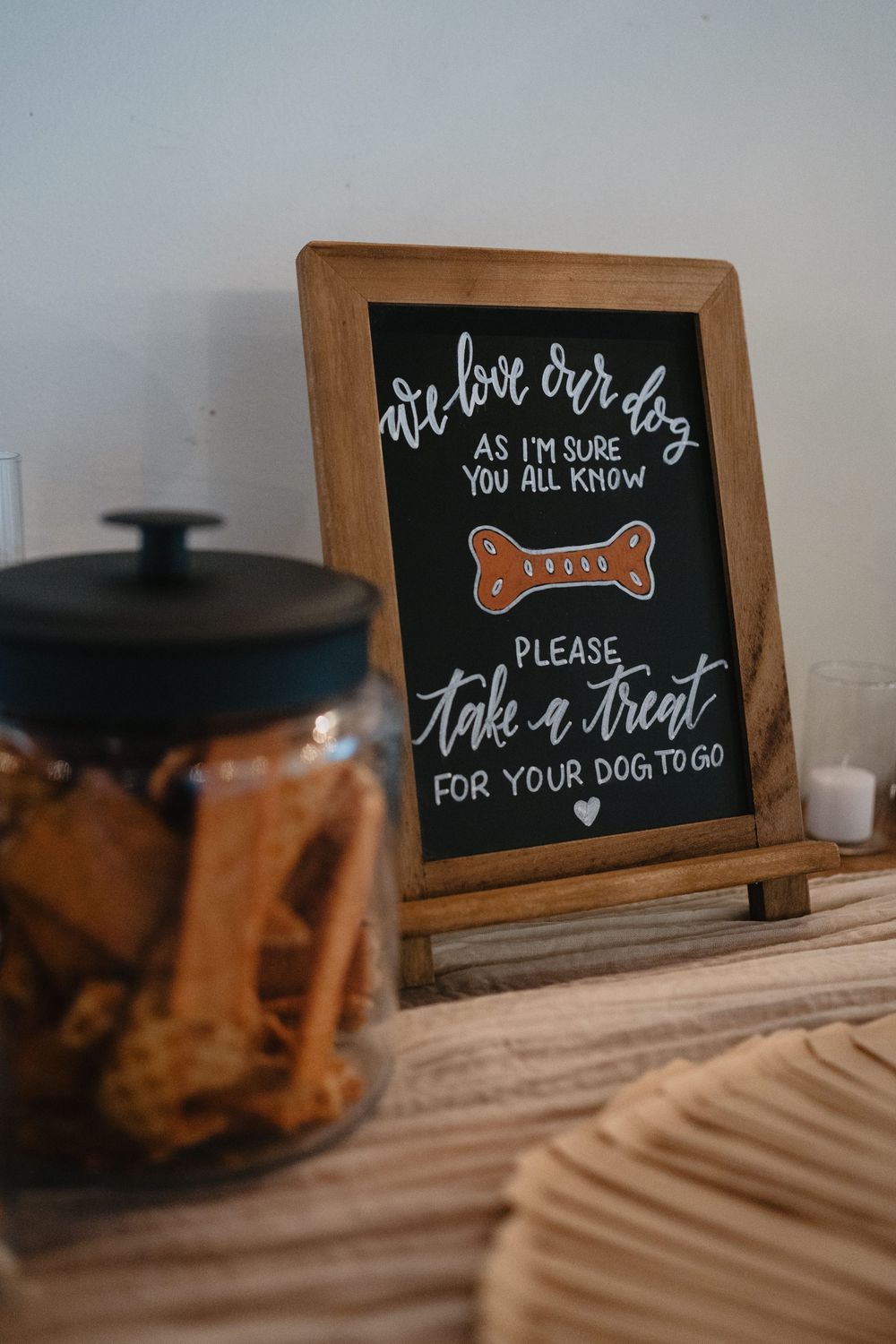 Chalkboard sign inviting wedding guests to take dog treats from a jar, displayed on a wooden table.