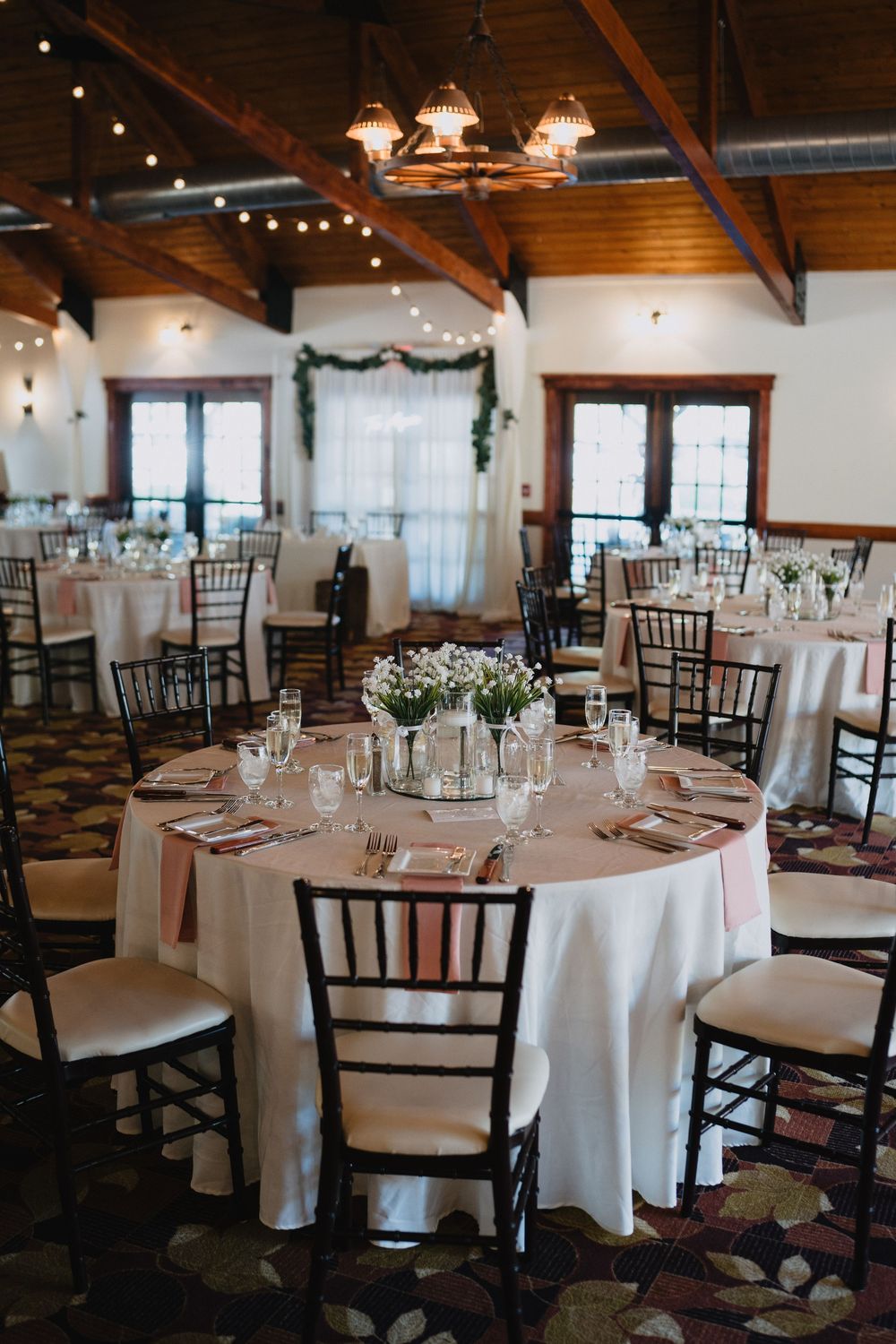 Elegant wedding reception hall with round tables, white linens, chiavari chairs, string lights, and floral centerpieces.