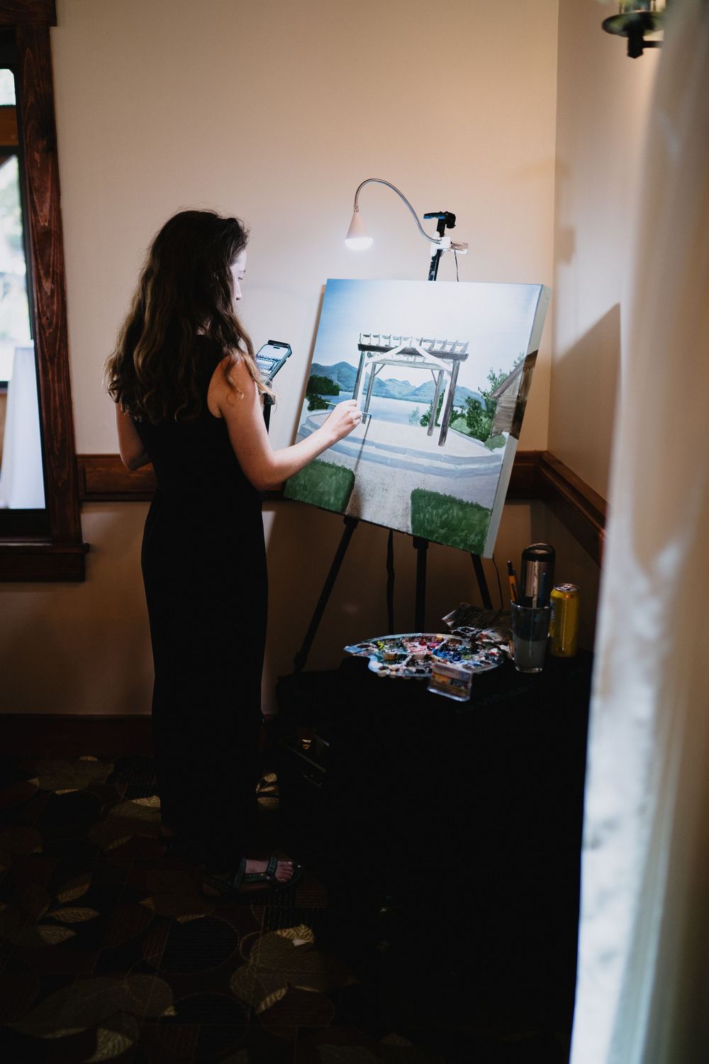 A woman in a black dress paints a wedding arch scene on canvas at an easel during a live painting event.