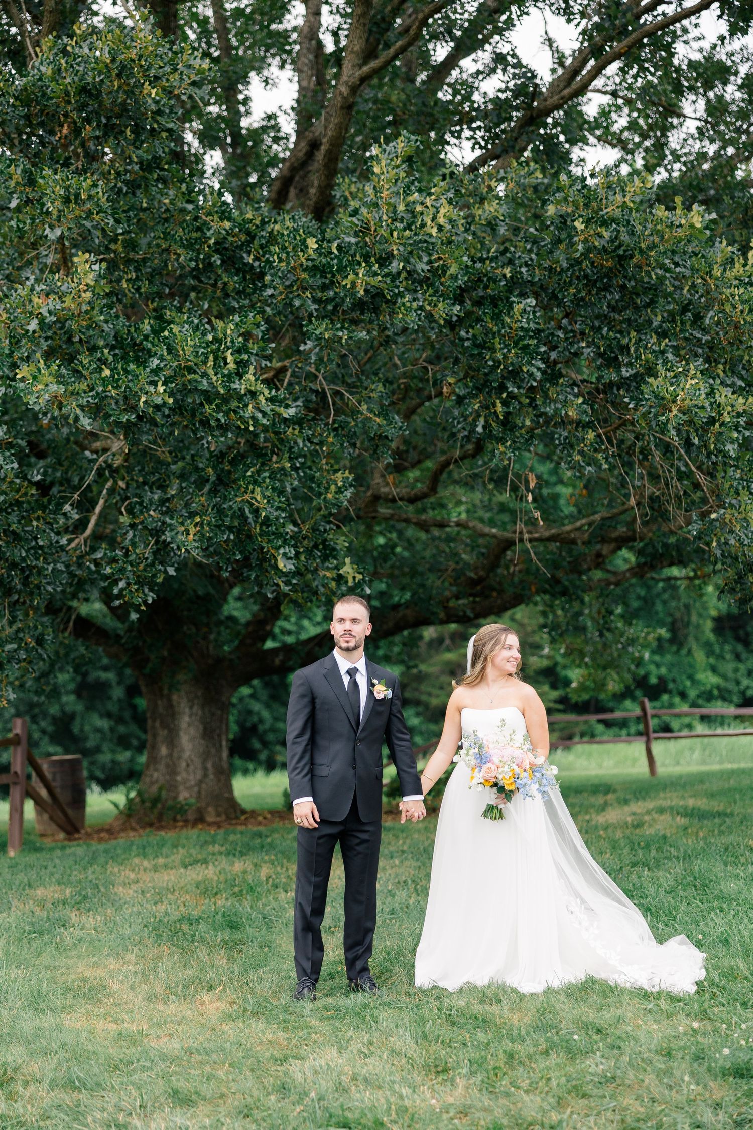Bride and groom smiling together outdoors, she holds a white floral bouquet, he wears a black suit with tie.