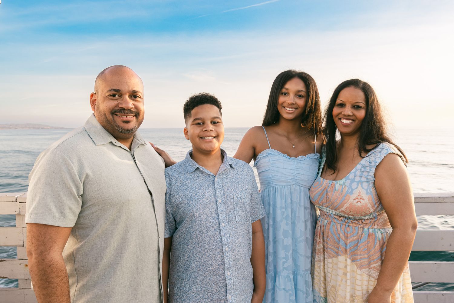 Family beach photos of a smiling family of four standing together on a pier overlooking the ocean at sunset.