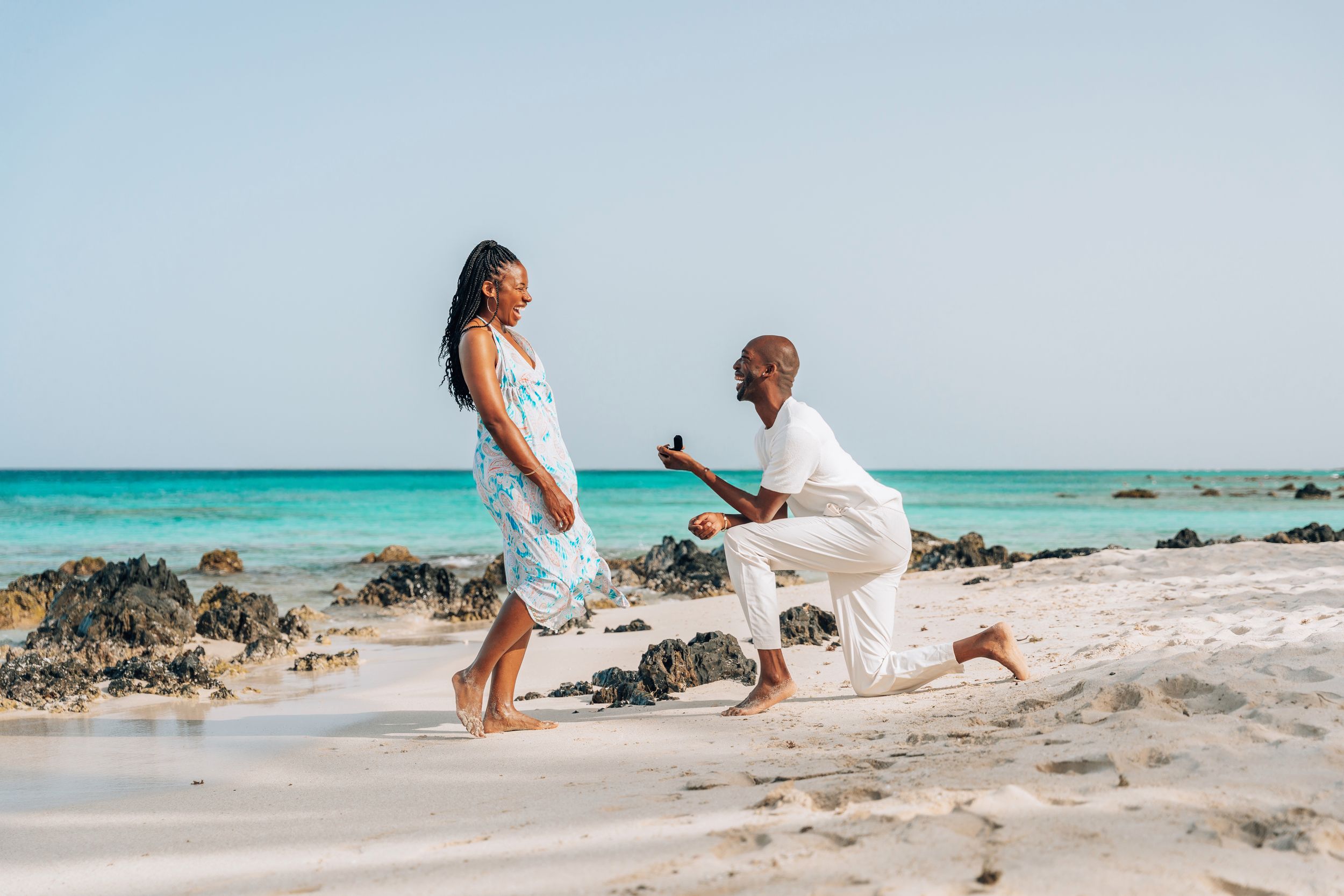 Couple holding hands at sunset beach proposal setup with floral arch, candles, and Marry Me sign on sandy shore.