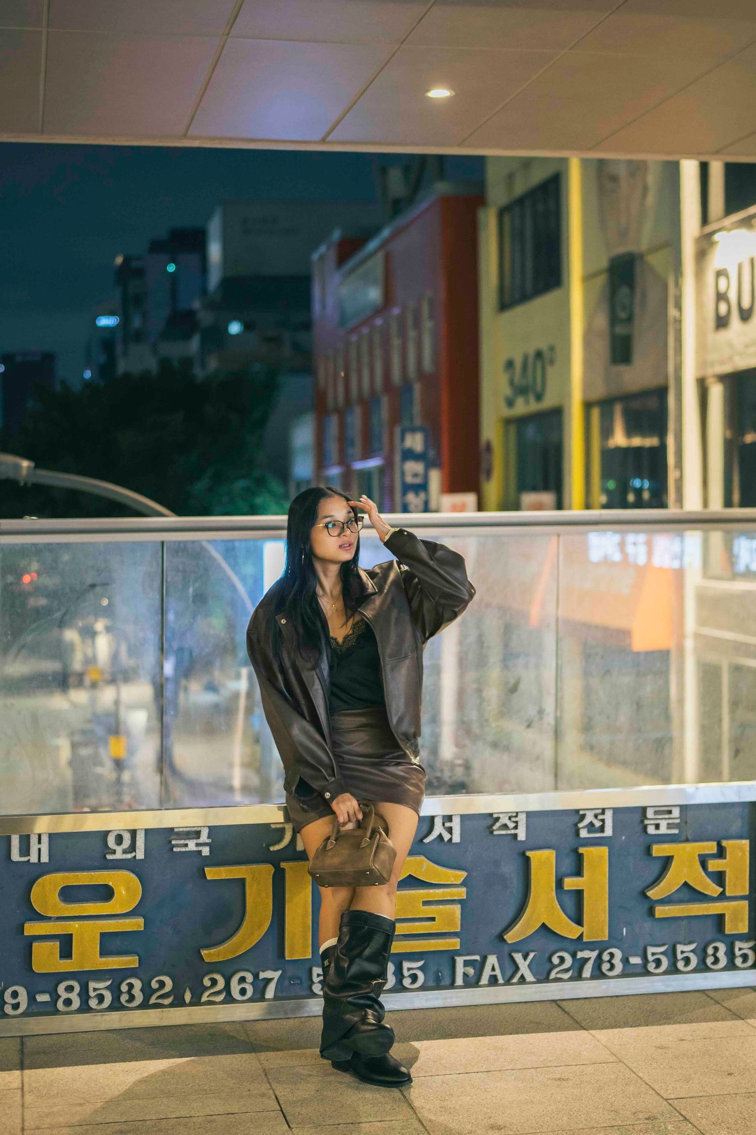 Woman in leather jacket and mini skirt posing at night outside a Korean street storefront.