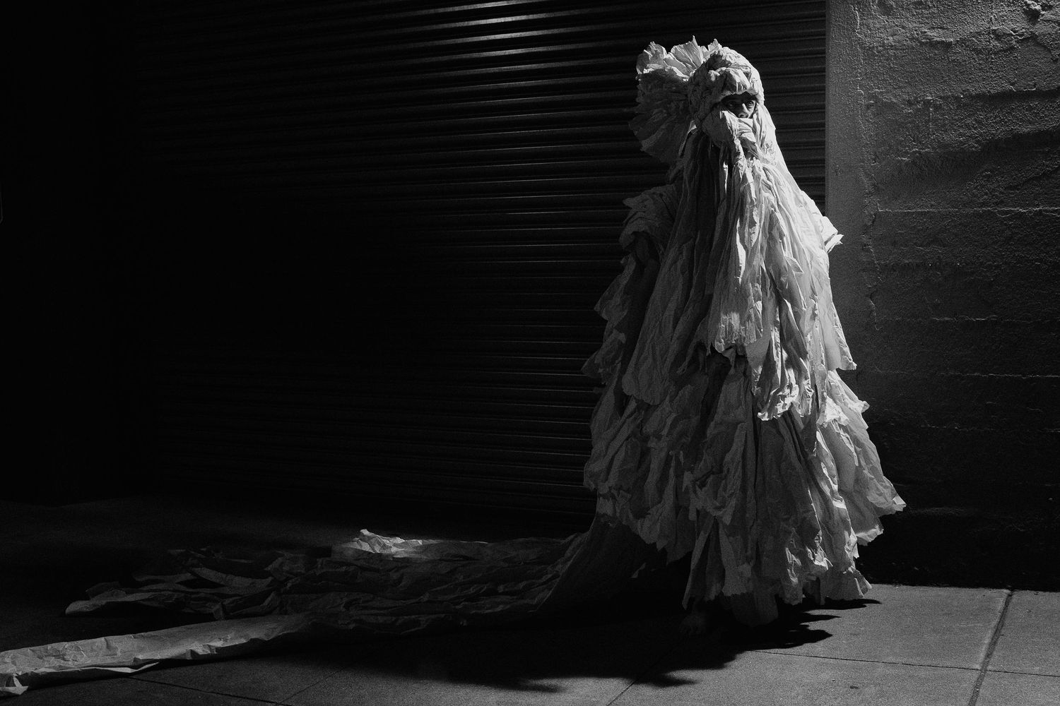 Black and white photo of a figure in an elaborate white ruffled gown with long train in dramatic dark setting.