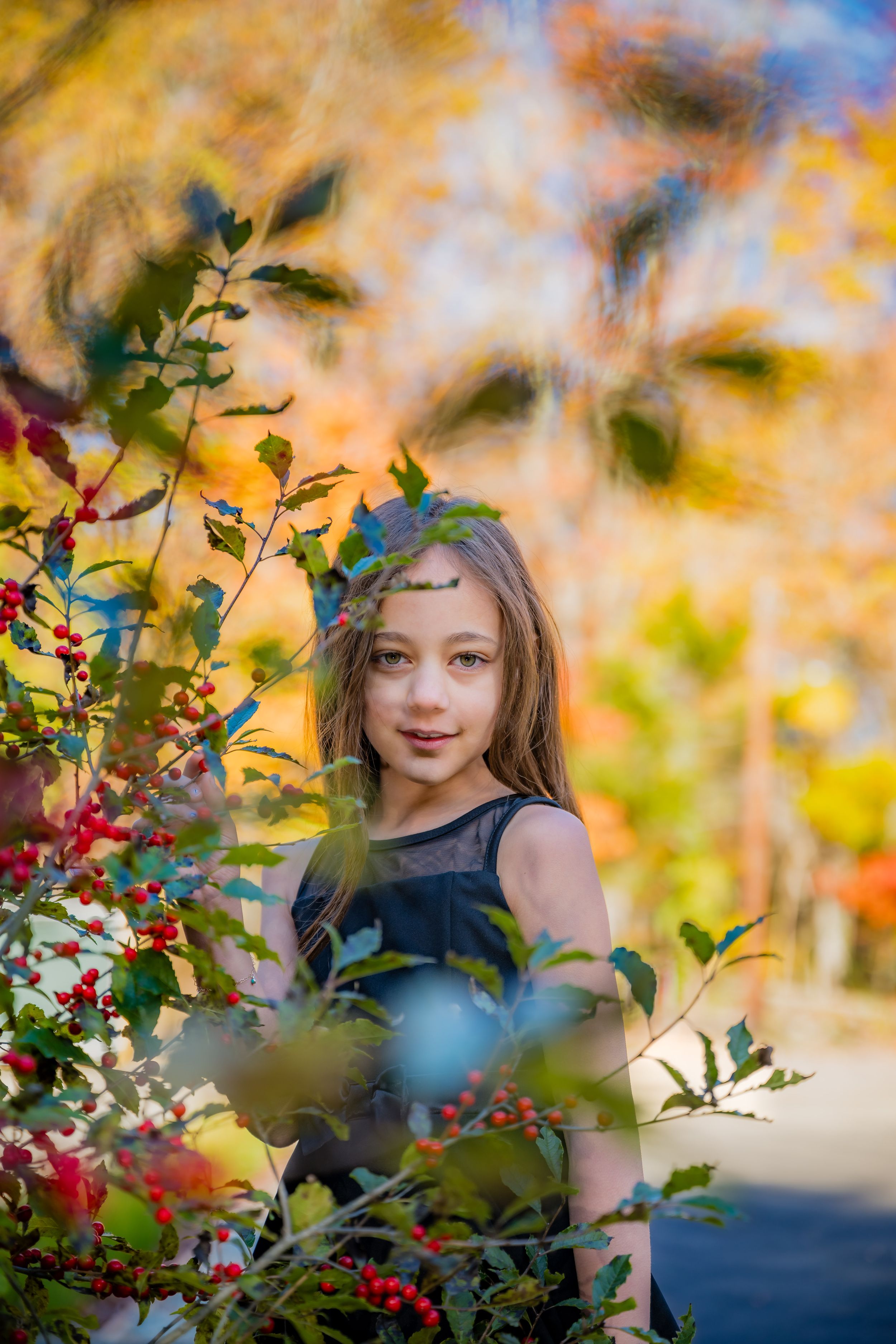 Young girl in black dress poses among red berry branches with colorful autumn foliage in background.
