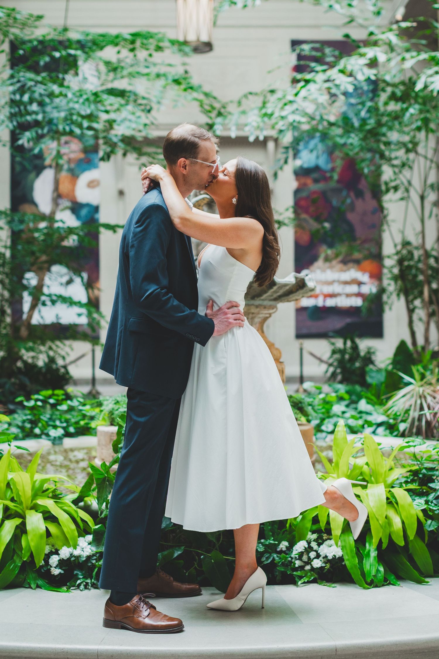 Bride and groom share a romantic kiss in a lush indoor garden, bride wearing a white tea-length dress and heels.