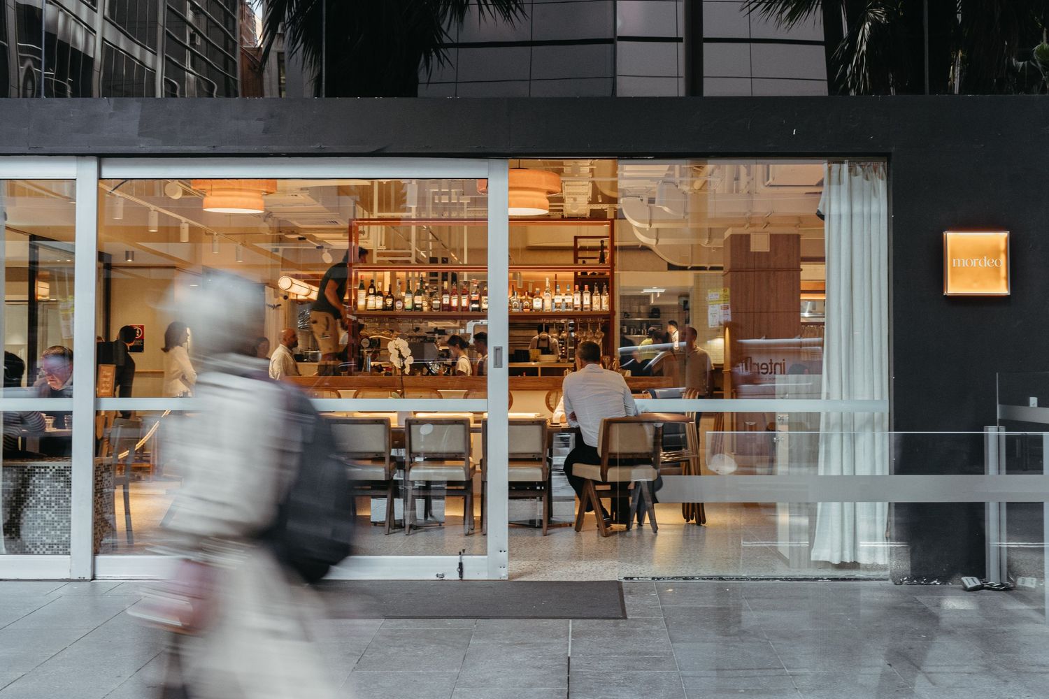 Person walking past a warmly lit modern restaurant with large glass windows at dusk.
